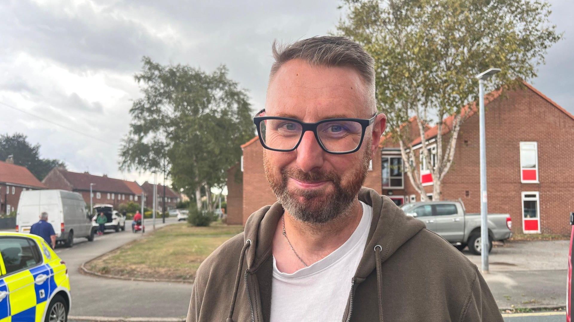 A head and shoulders shot of a man smiling at the camera. He has short brown hair and is wearing glasses, a white T-shirt and a khaki zip up hoodie. The back of a police car can be seen on the left. Houses are in the background.