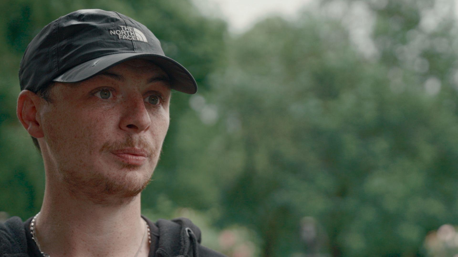 Close-up portrait of Darren Docherty wearing a black baseball cap outdoors. The image is focused on his face, with a softly blurred background of green trees and foliage.