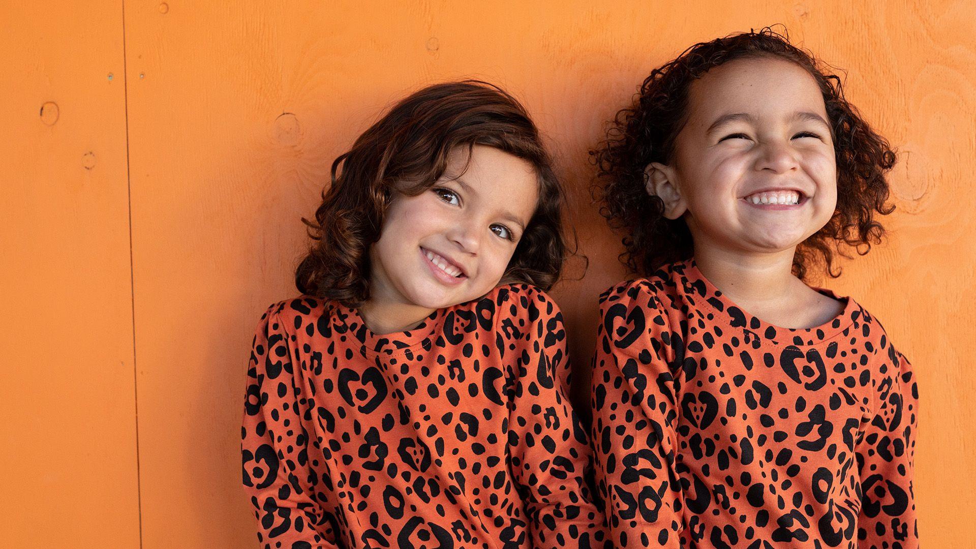 Twin girls with brown curly hair and orange leopard print tops laugh with each other