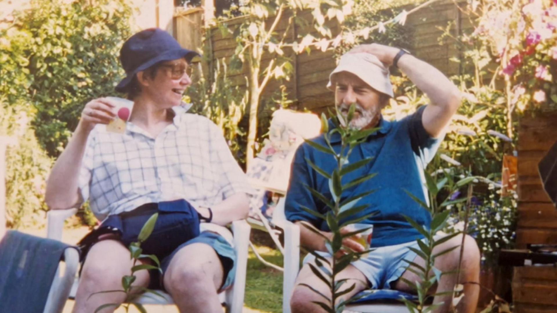 Gillian and Les Holmes sat in in a garden on deckchairs on a sunny day. They are holding drinks and wearing sun hats