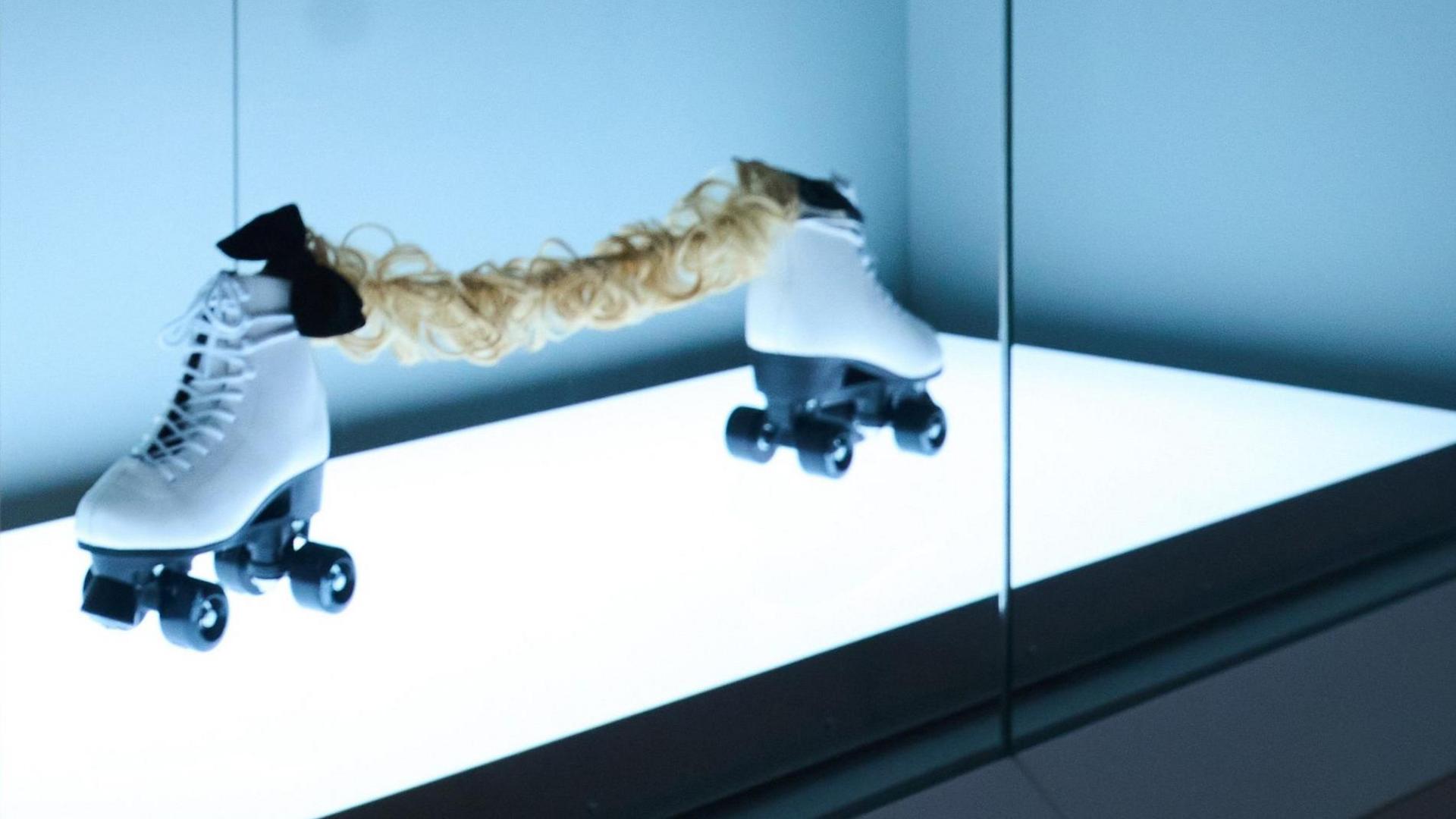 An art exhibit behind glass, which is lit from below. A pair of white roller skates with black wheels sit facing away from each other, with about 0.5 metres gap between them. They are joined by a curly blonde hair extension and two black bow ties.