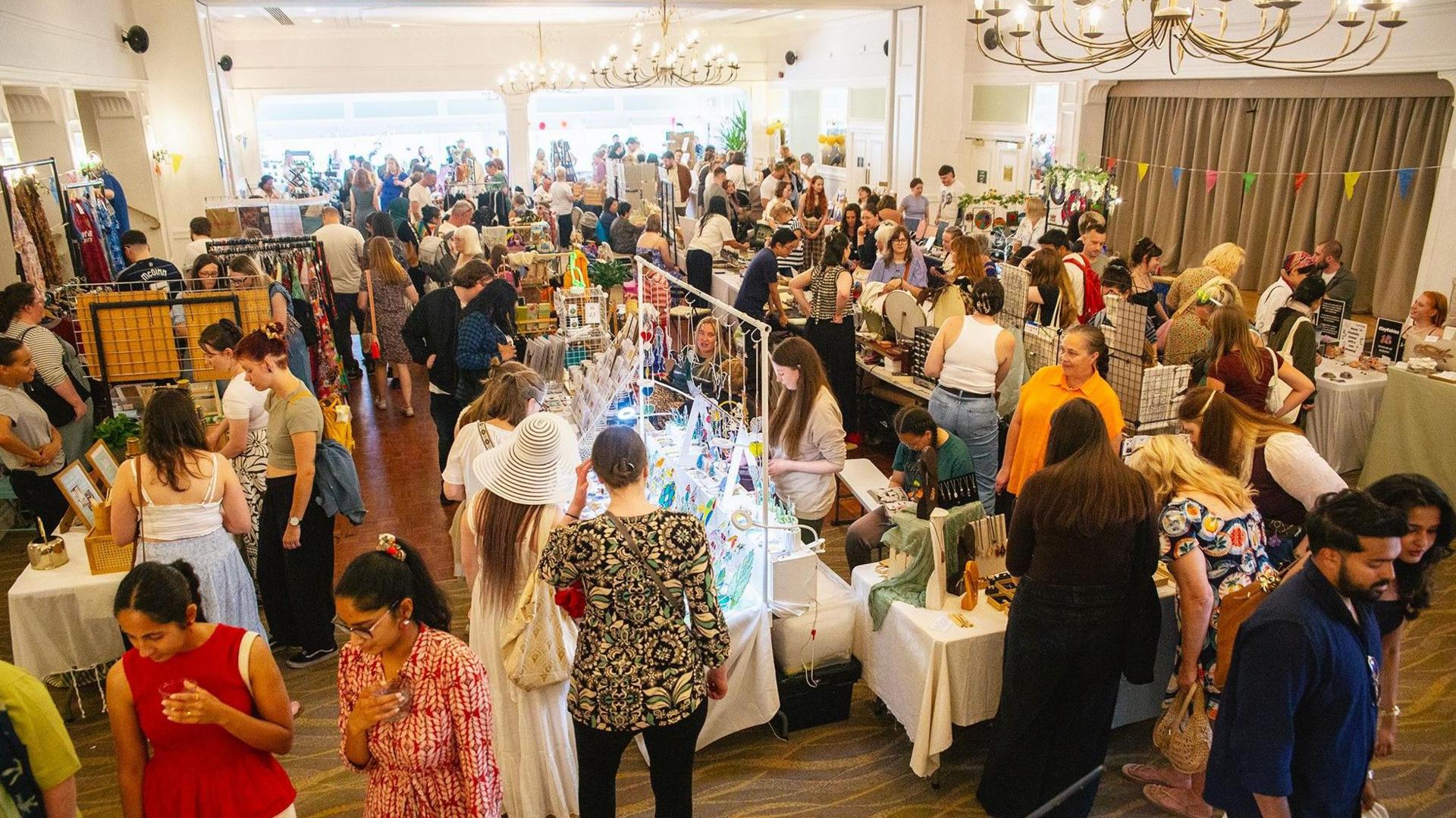 Dozens of people are looking at stalls inside a building. Scarves and wind chimes are among the items which can be seen. 