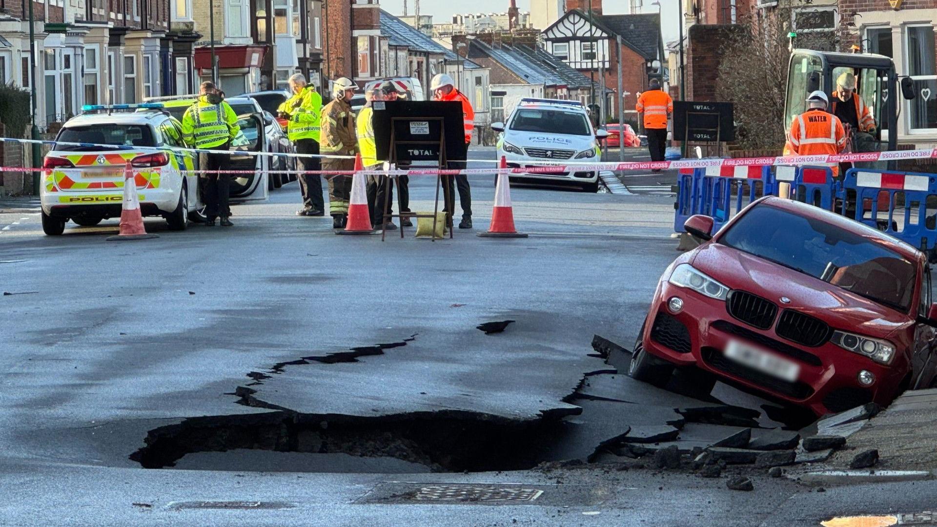 A red BMW slumped into the side of a road. Holes and cracks in the road surround the car. A police cordon is in place behind it with various emergency service workers gathered.