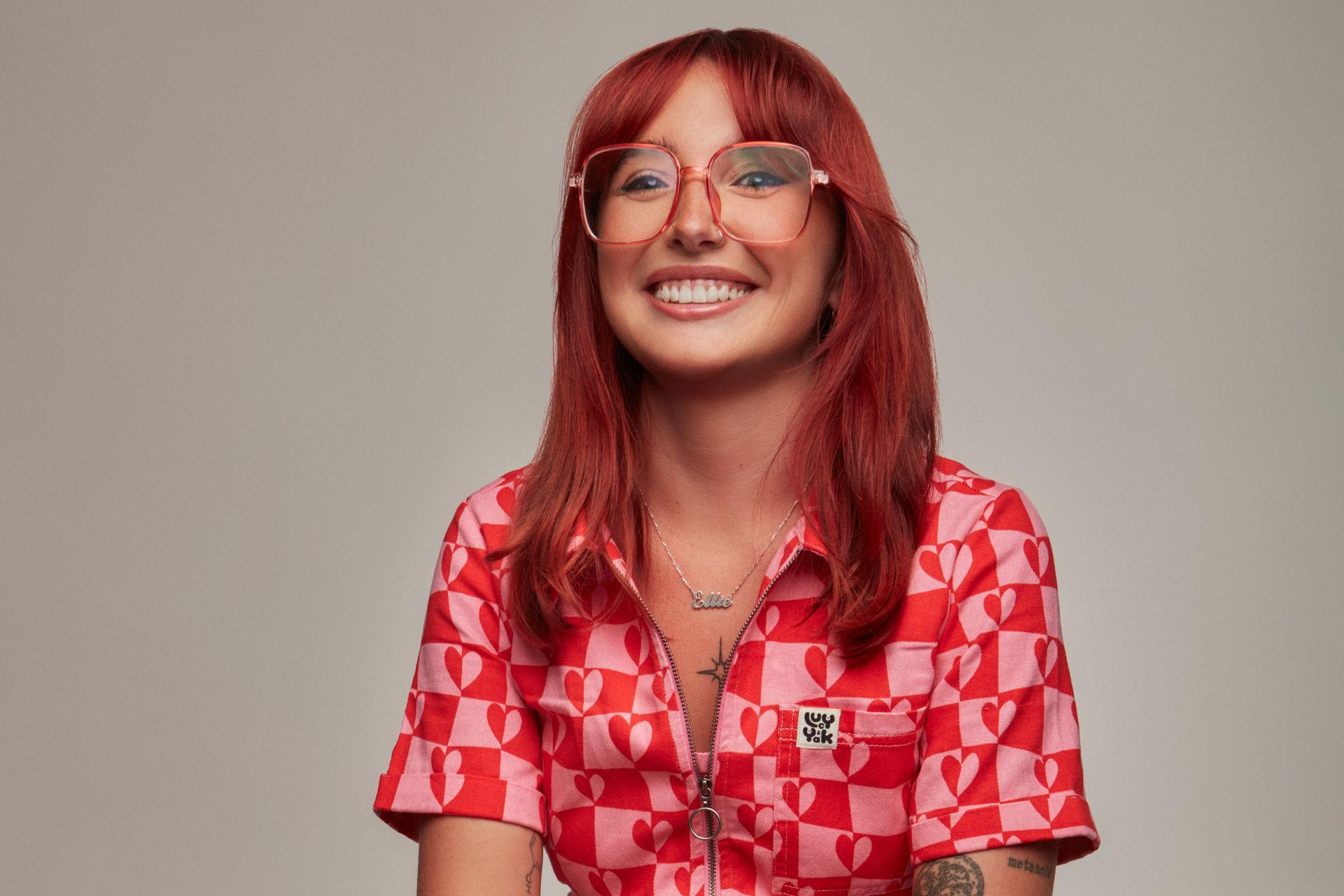 Ellie Middleton, who has long, red hair and is wearing glasses, is smiling against a plain grey backdrop