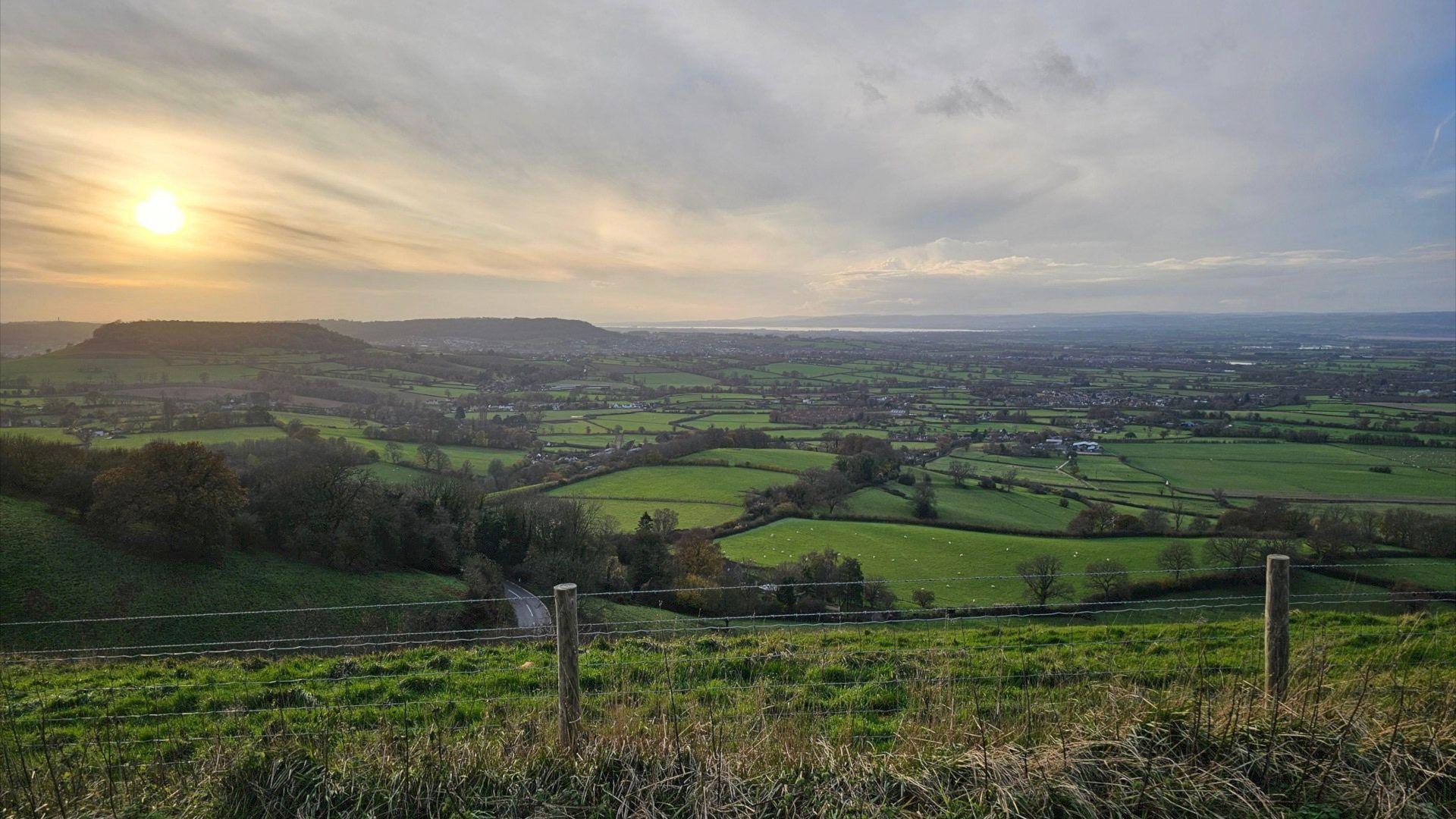 Sunrise at Coaley overlooking a spread of fields with hills visible in the distance and multiple green fields bordered by dark hedges in the foreground