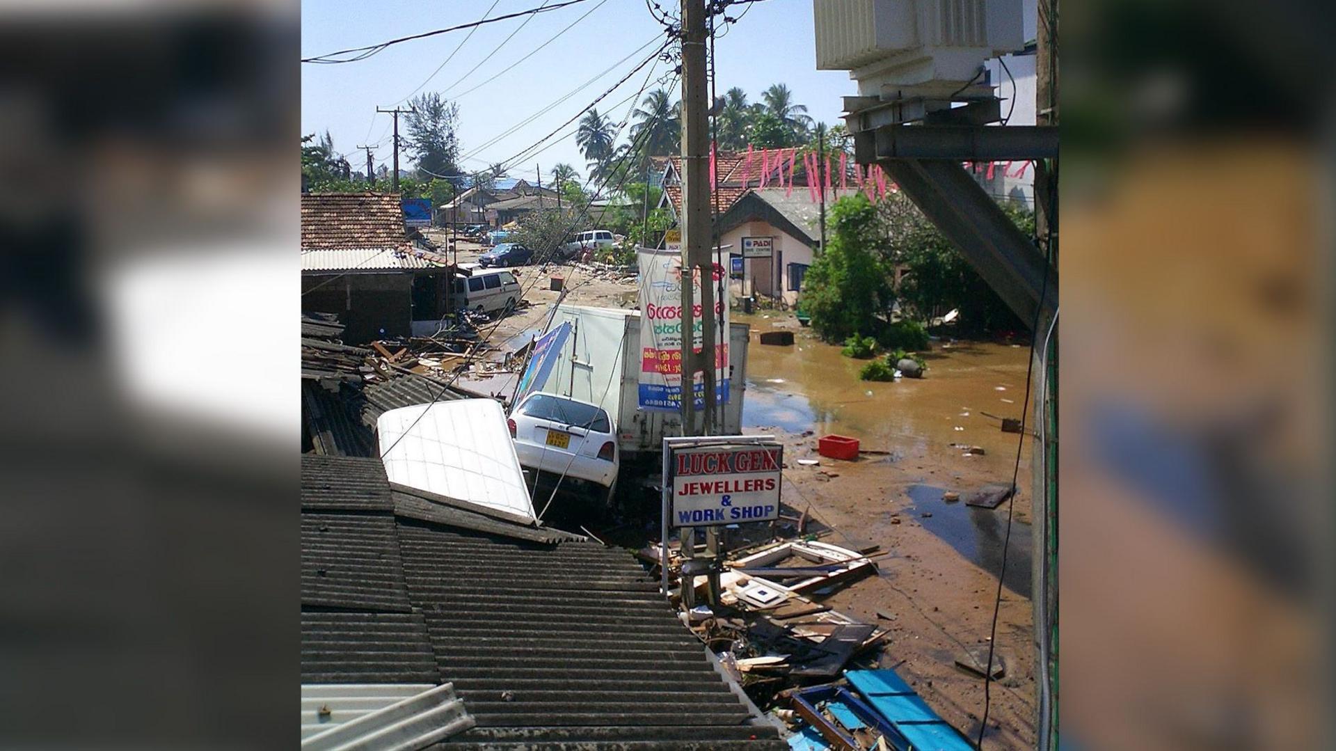 A street, devastated by flooding. It is filled with muddy water and collapsed buildings.