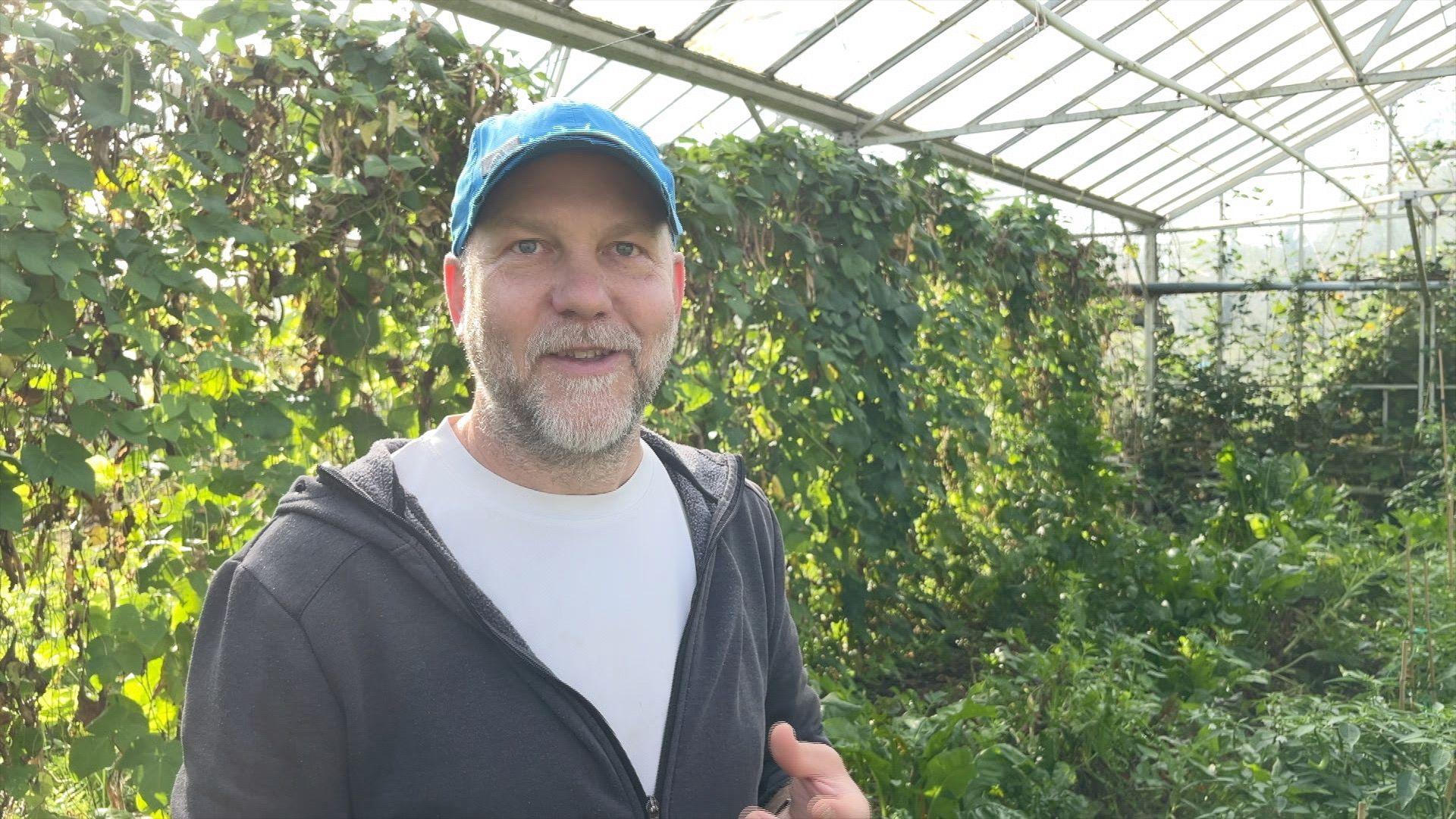 A man with a beard and a blue hat in a white T-shirt and jumper stands in a glass house surrounded by green vines.