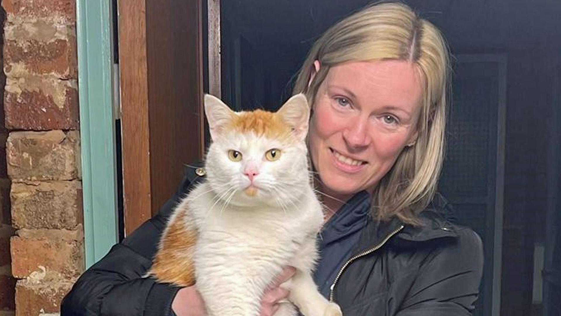 Cara McBurnie holds Beans the cat - both look into the camera. Cara is blonde, with blue eyes and is smiling, wearing a black jacket and dark coloured top underneath. Beans is mainly white, with ginger patches and yellow eyes. They are standing in a doorway with a green trim around the door and bricks to the side.