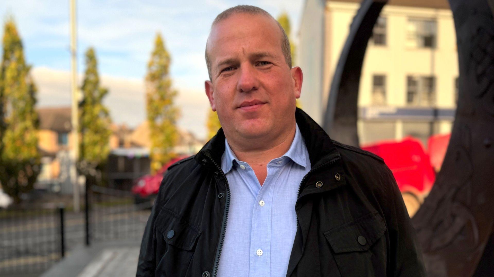 Clement Cuthbertson looks into the camera on a town street in the daytime. He is wearing a black jacket and blue shirt. 