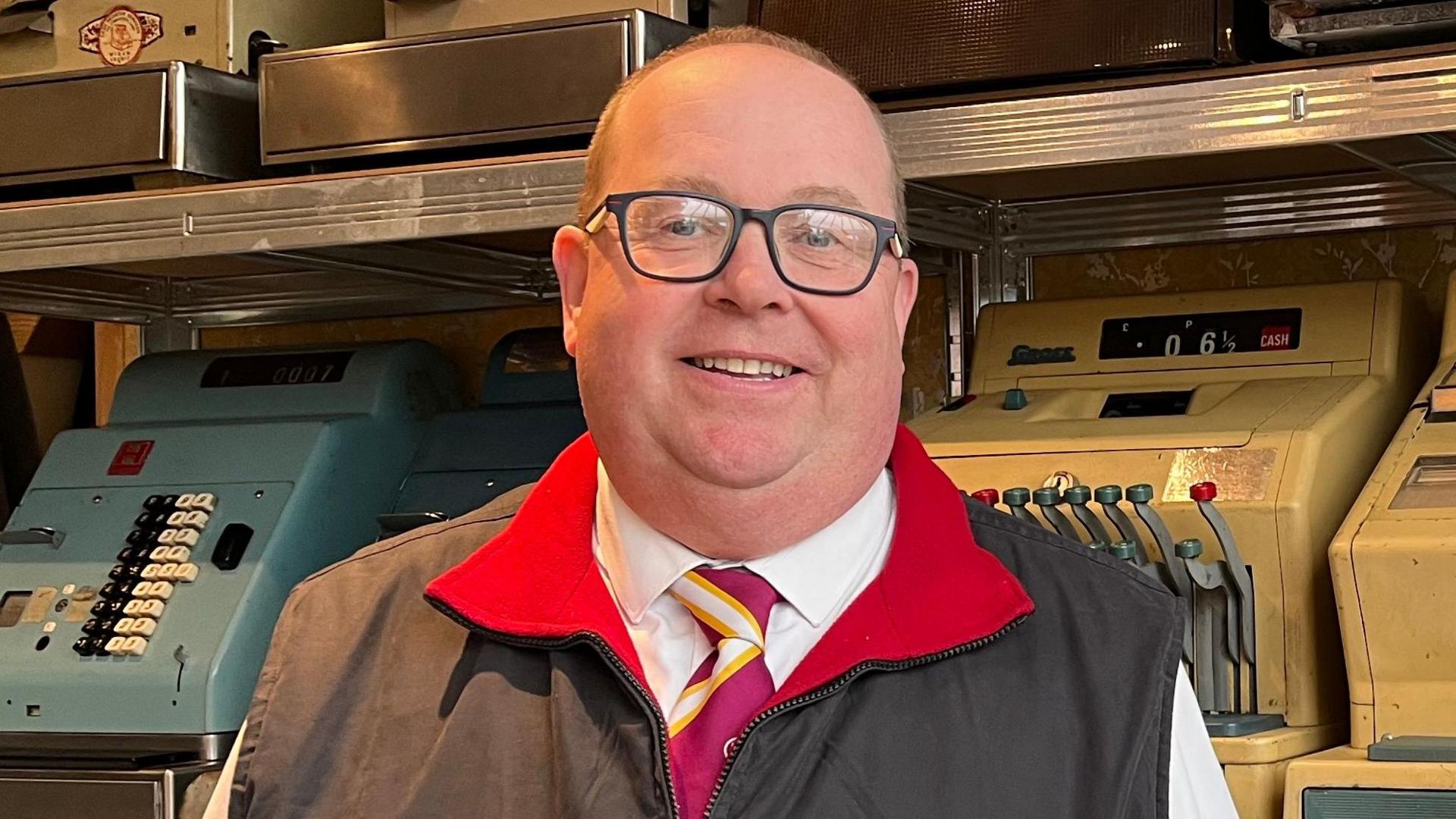 Andrew Rae is wearing a black jacket with a red collar, he has a purple and yellow tie and is standing in front of his cash register collection.