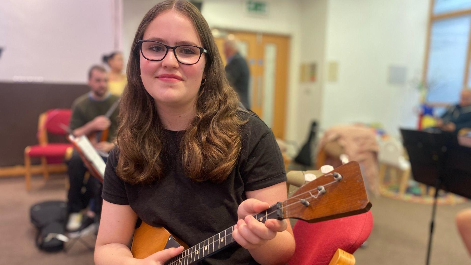 Isla Graham, with long brown hair and wearing a black t-shirt and black framed glasses, poses with her balalaika.