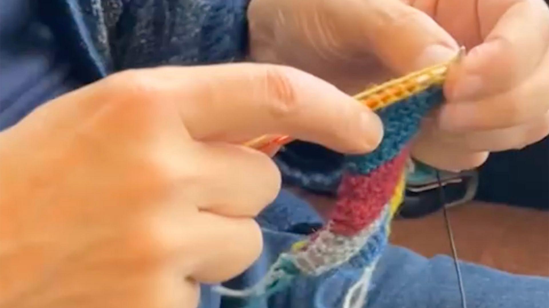 Man's hands knitting segments of a yellow, blue, red and grey wool hat
