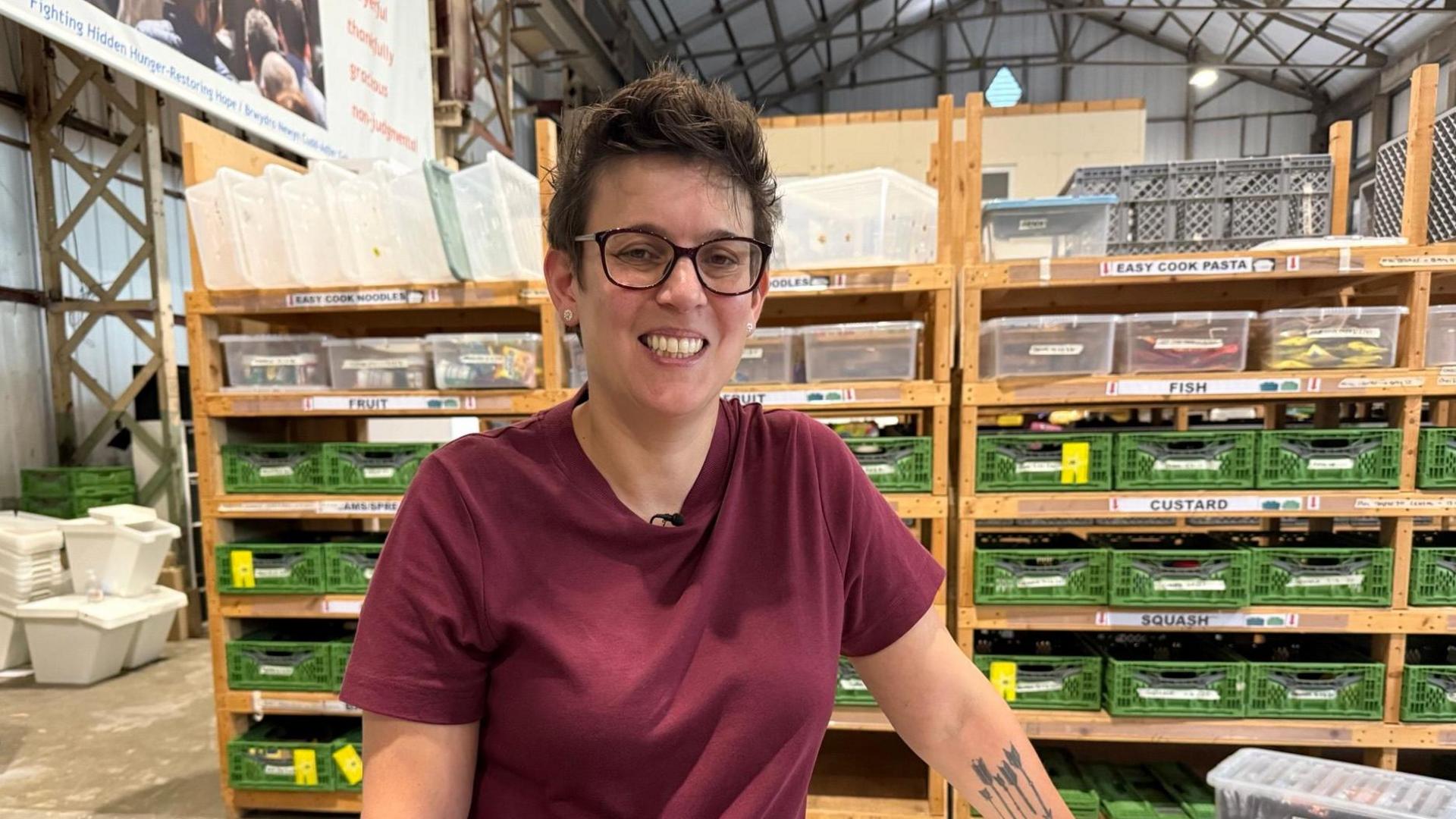 Rachel Biggs, CEO of Cardiff Foodbank wears a purple T shirt and has rectangular glasses and is smiling into the camera and has short curly hair