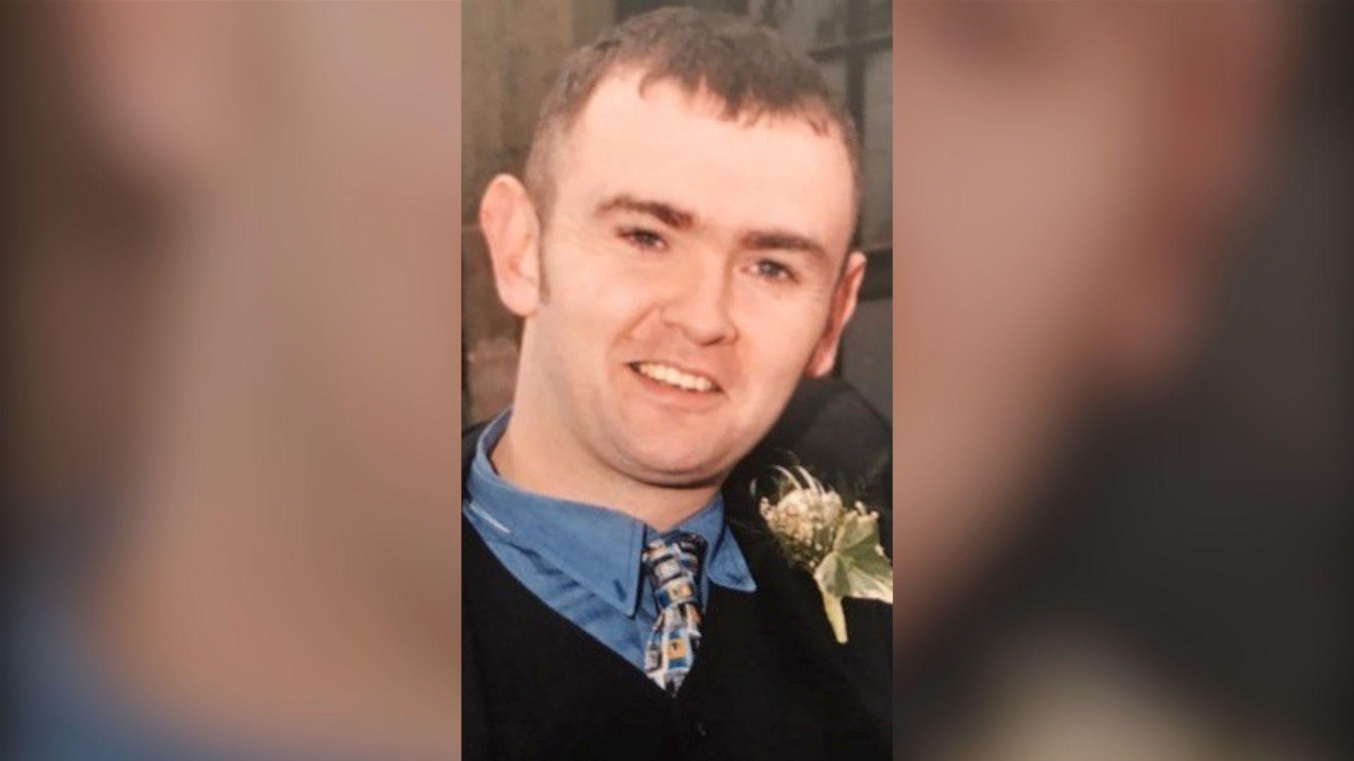 Mr Heagney with short dark hair, smiling at the camera as he wears a black suit, blue shirt and multicoloured tie.