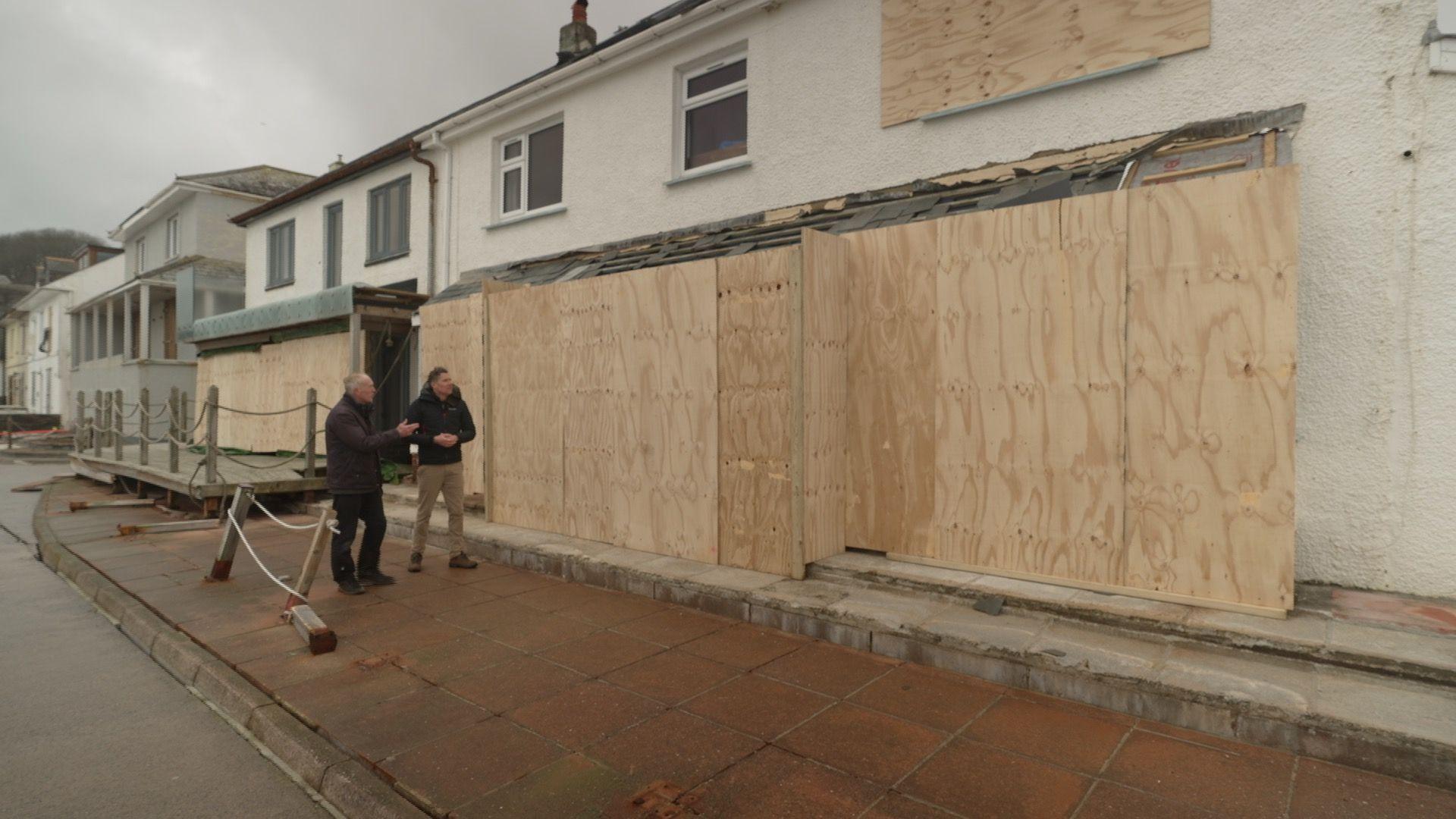 The image shows seafront properties in Torcross, Devon.  Several of the homes have windows and doors boarded up after being damaged in Storm Ingrid.  