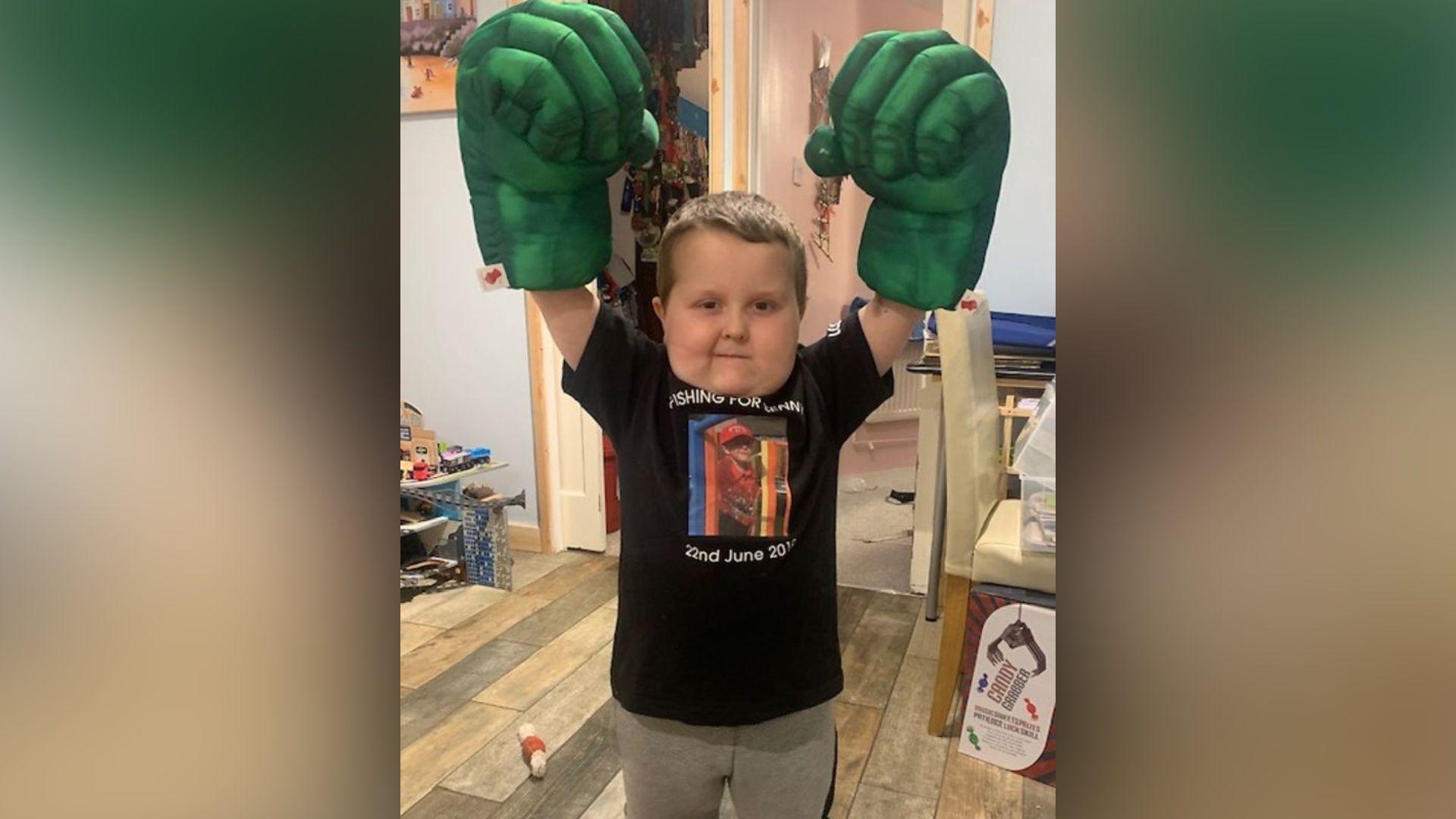Benny Pitcher wears huge green Hulk-style boxing gloves. he has his arms raised and is standing in a room with toys around him.