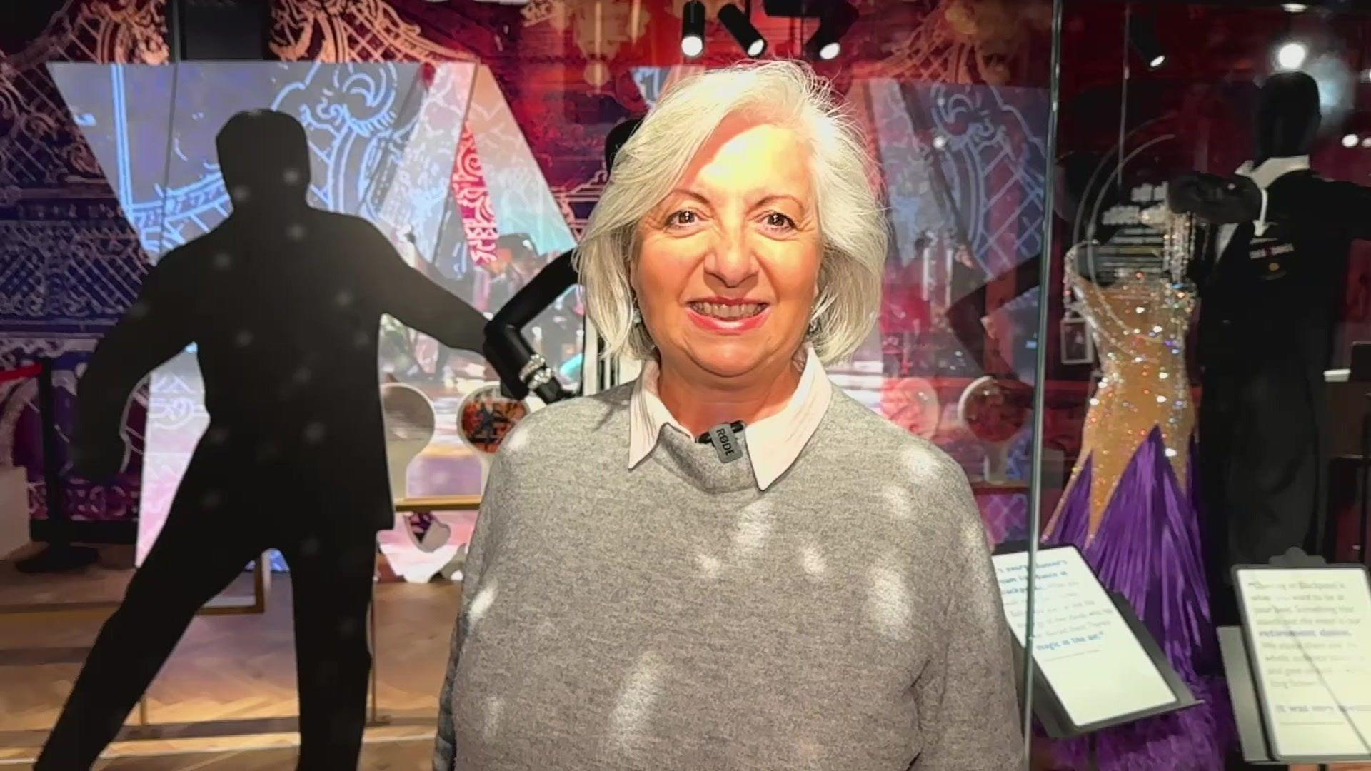 Marina Blore has bobbed length silver hair and wears a white blouse and grey jumper. She is smiling and is standing in front of the dance display and mannequins with costumes at Showtown.