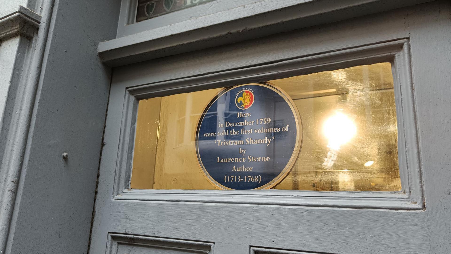 An image of a blue plaque in a window above a door. It says 'Here in December 1759 were sold the first volumes of 'Tristram Shandy' by Laurence Sterne Author (1713-1768)