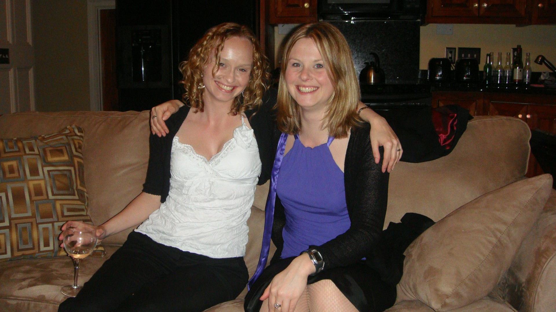 Two women sit on a brown sofa with their arms around each other. They are smiling at the camera. One is holding a glass of white wine in her right hand.