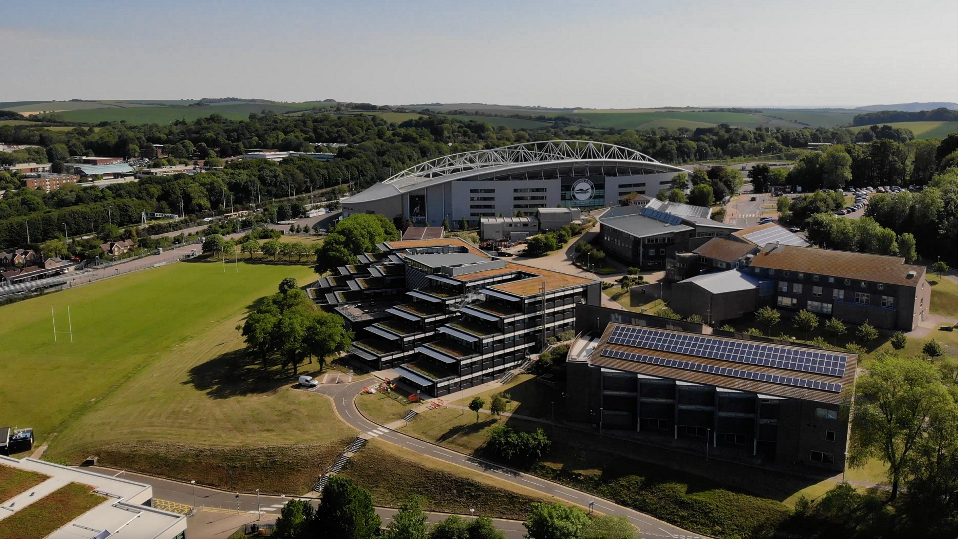 An aerial view of the university's Falmer campus which shows a modern building in the background with a white metal arch reaching over the top of it and in the foreground stepped buildings and brick buildings with solar panels on the roof. There are also sports pitches in the picture and green hills in the background.