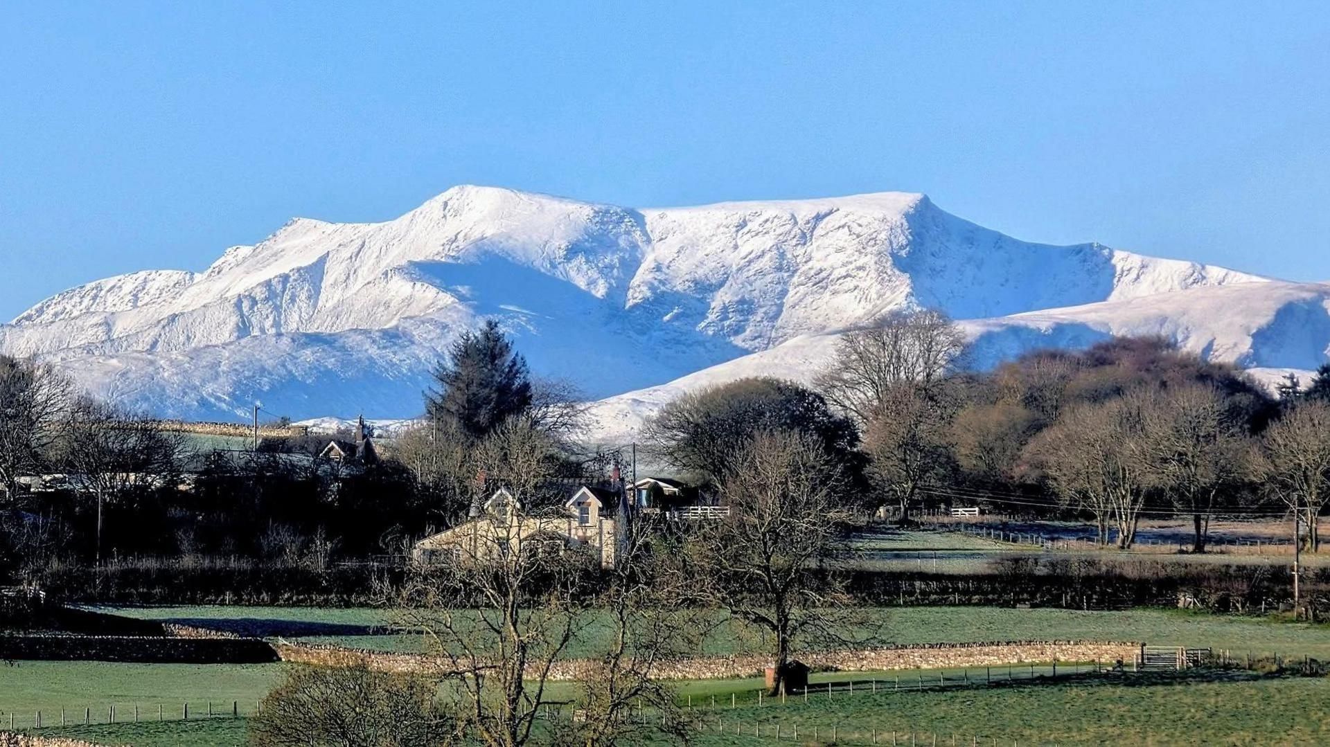 Blencathra mountain in the Lake District. Snow has cloaked the mountain and it has blue hues in reflection from the sky which is also bright blue. There is a farm house in the foreground and green grass.