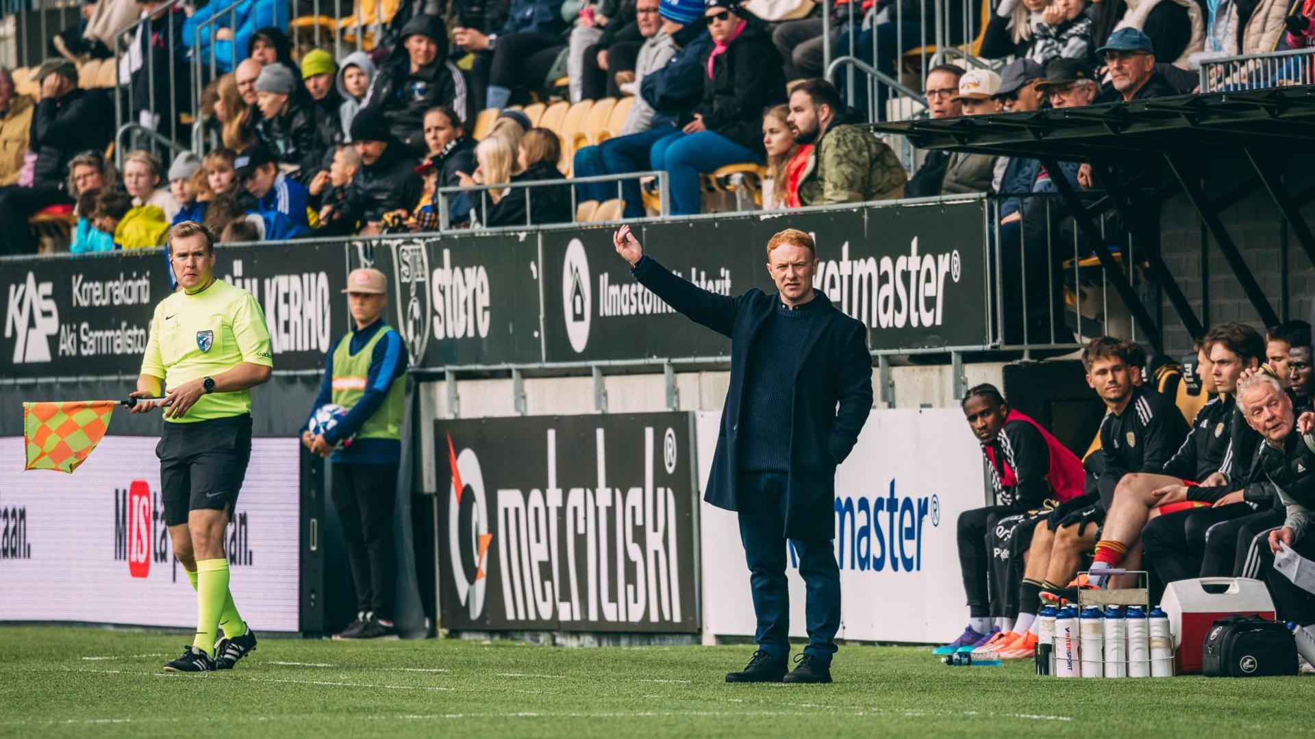 SJK Seinajoki manager Stevie Grieve stands with his arm aloft in his technical area, as fans and players look on around him.