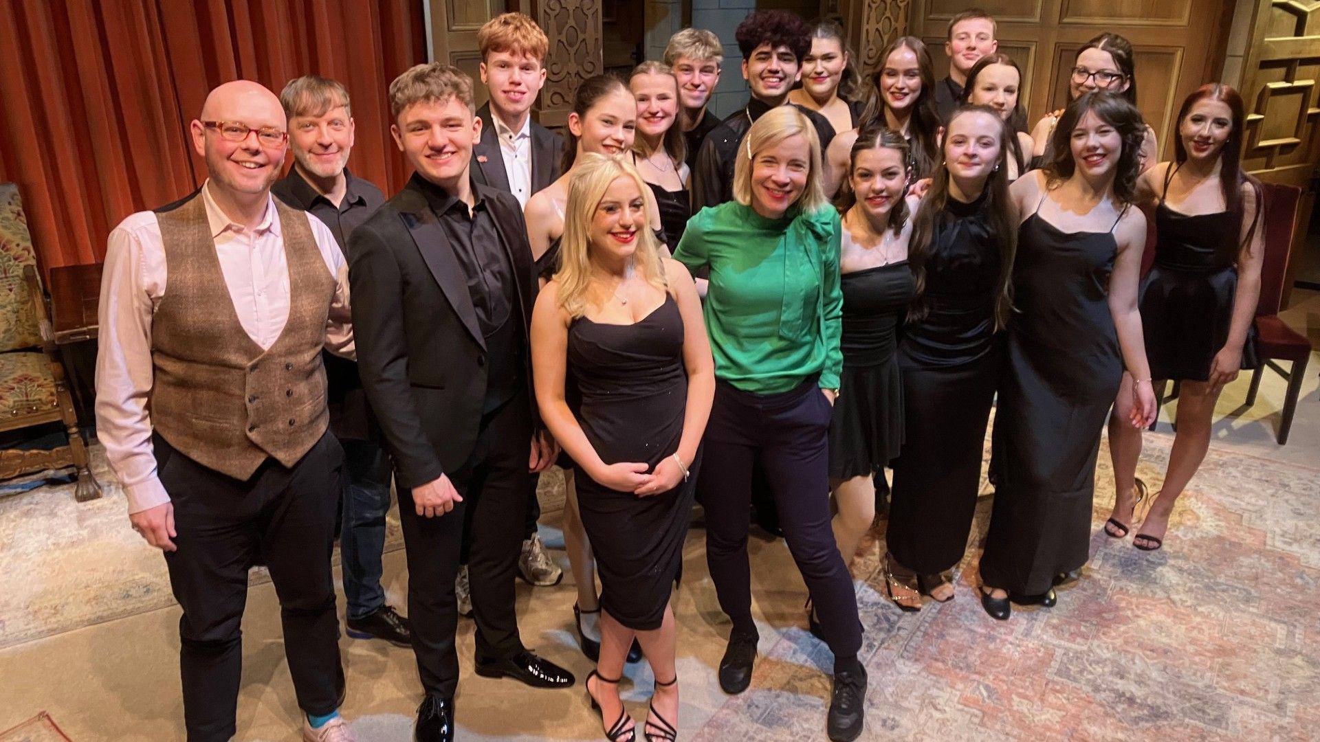 A crowd of teenagers stand with their teacher, Ian backstage at a theatre, all wearing smart dresses and suits, smiling at the camera