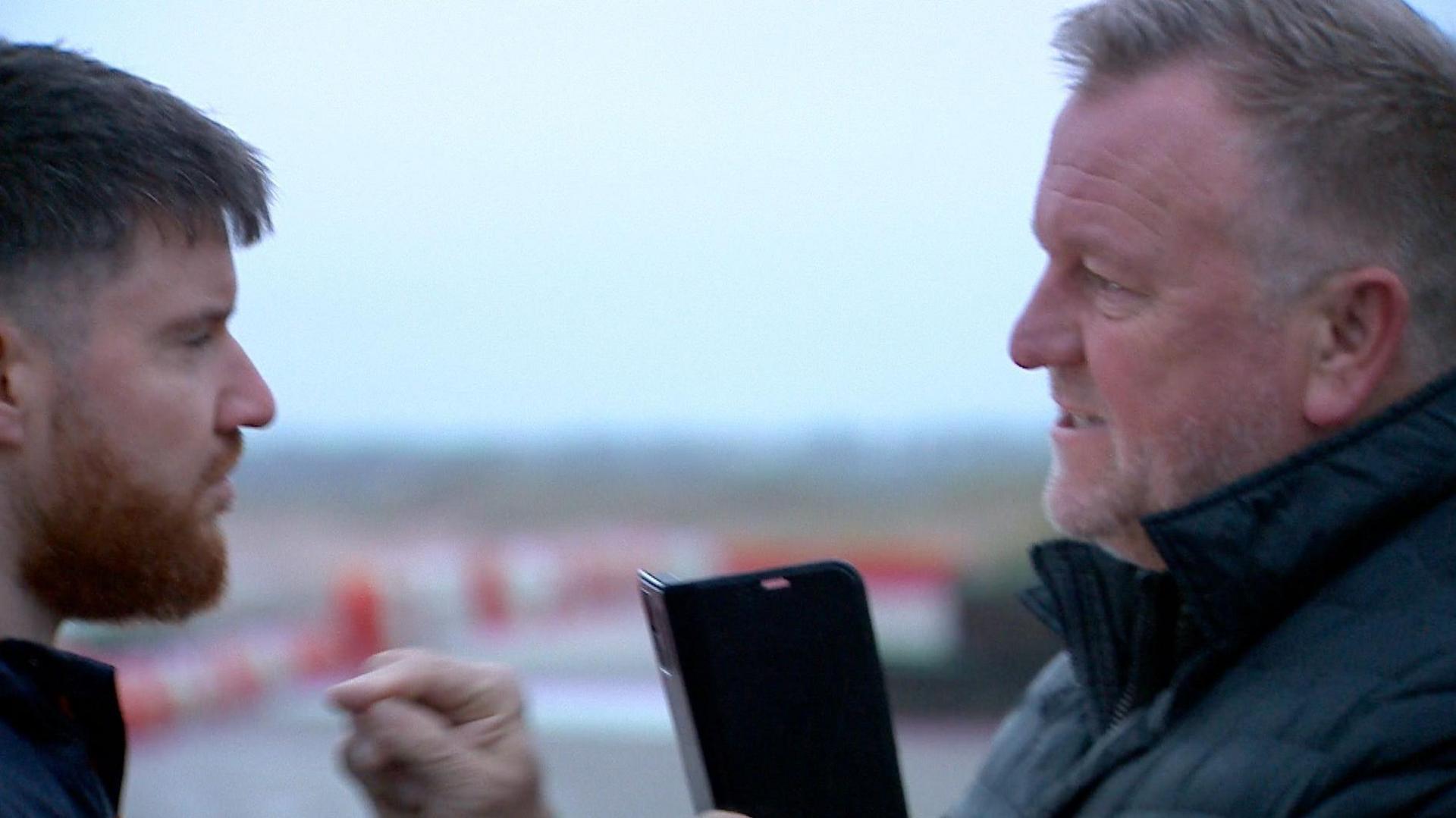 Barry Elliott has short greying hair, combed forwards from the temples, with a slight peak at the forehead and a grizzled white grey beard and slight moustache. He is wearing a black coat with the collar up. He is mid sentence, with his mouth open, looks stern and is pointing a finger at a BBC reporter. He has his mobile phone in the other hand.