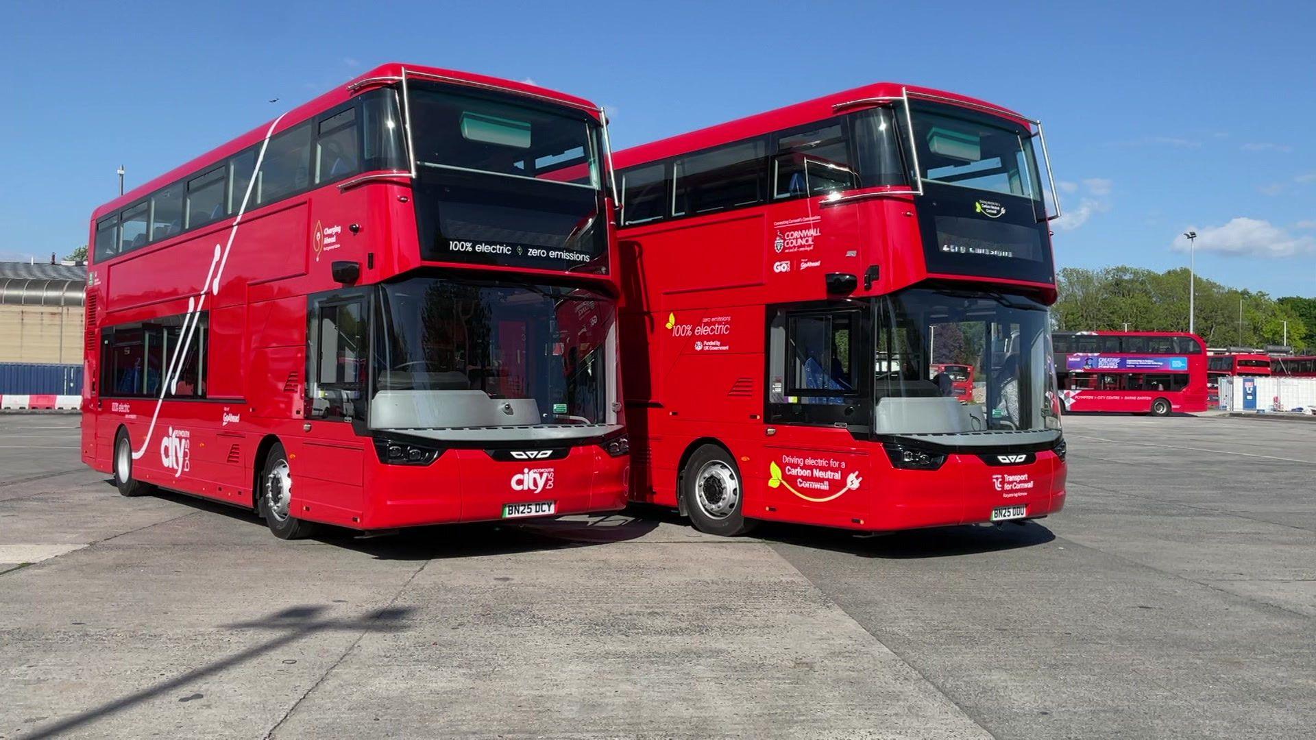 Two red double-decker buses are parked side by side in an open area. Both display the text '100% electric • zero emissions' on the front. The left bus has 'city' written on it, while the right bus includes branding for a project with Cornwall Council and partners. Additional red buses are visible in the background.