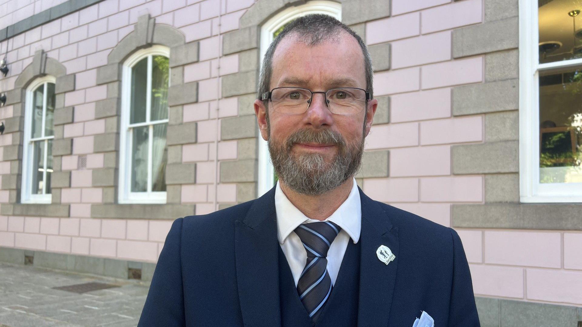 Deputy Malcolm Ferey is smiling at the camera as he stands on the pavement outside a pink granite building. He has short grey and brown hair and a beard and is wearing glasses. He is wearing a navy jacket and waistcoat over a white shirt and navy and pale blue striped tie. He has a silver pin badge and a pale blue handkerchief in his top pocket.