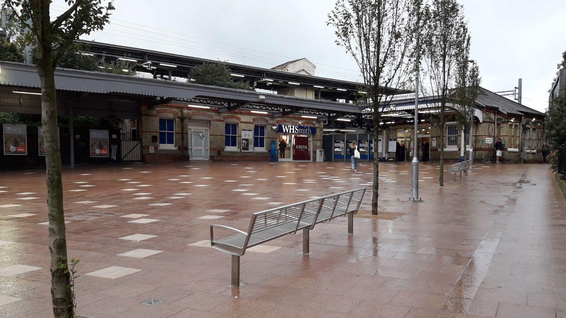 An exterior view of Maidenhead Station, the flooring is a red brick pavement, there are trees planted and benches in between them