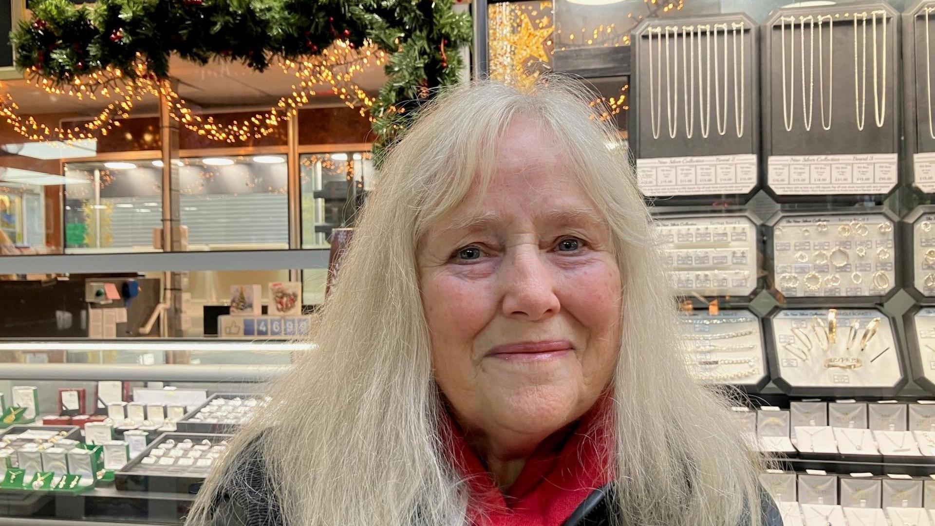 An older woman with long white hair standing next to a jewellery stall