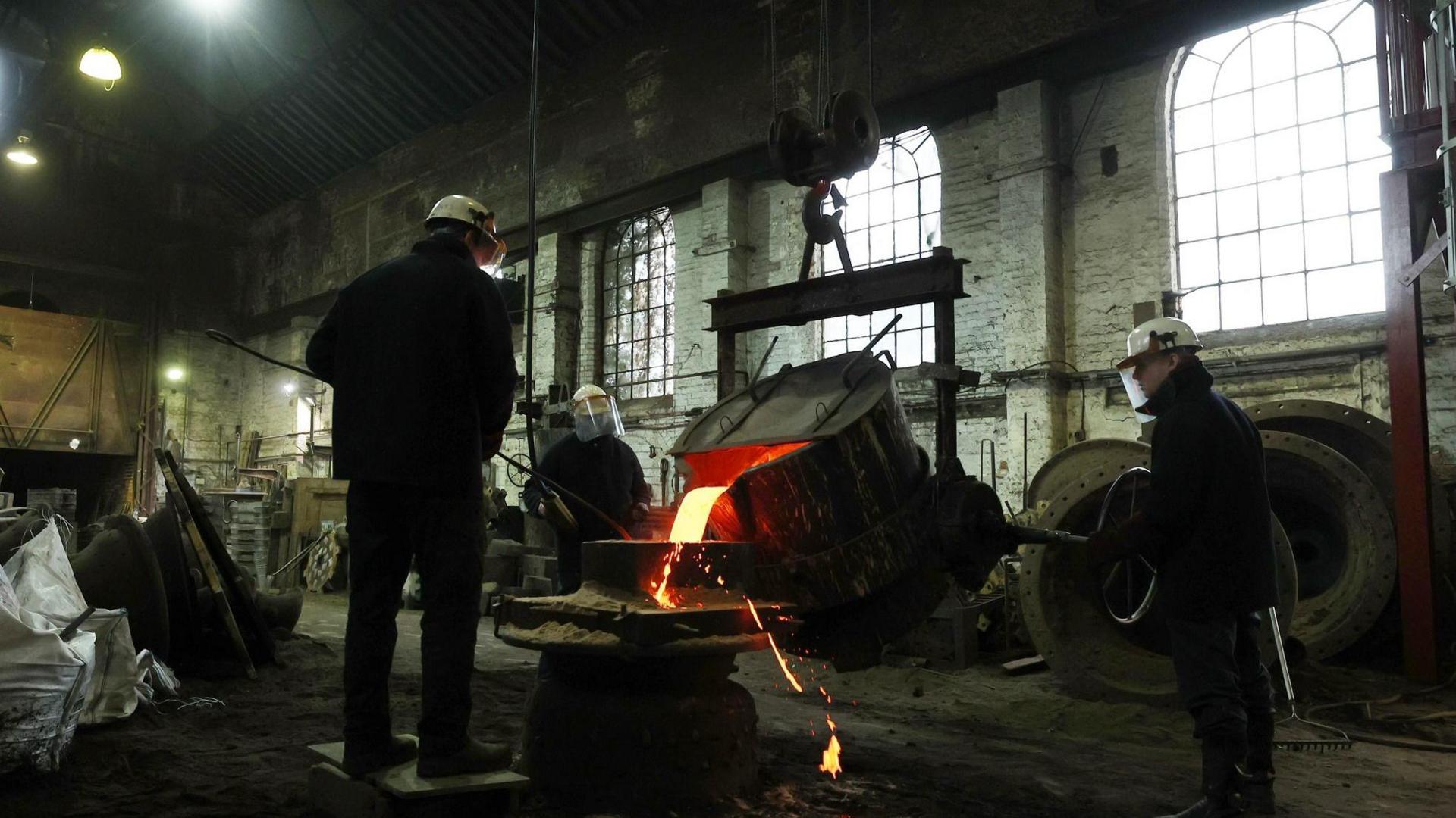Three workmen in protective clothing pour molten metal into a bell casting case
