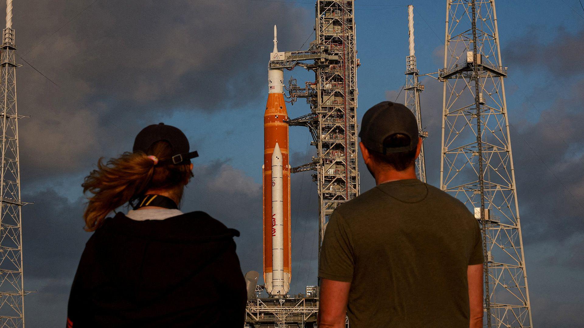 A woman on the left and a man on right hae their backs to the camera. They are looking at the Artemis rocket which is stood in place on the platform.