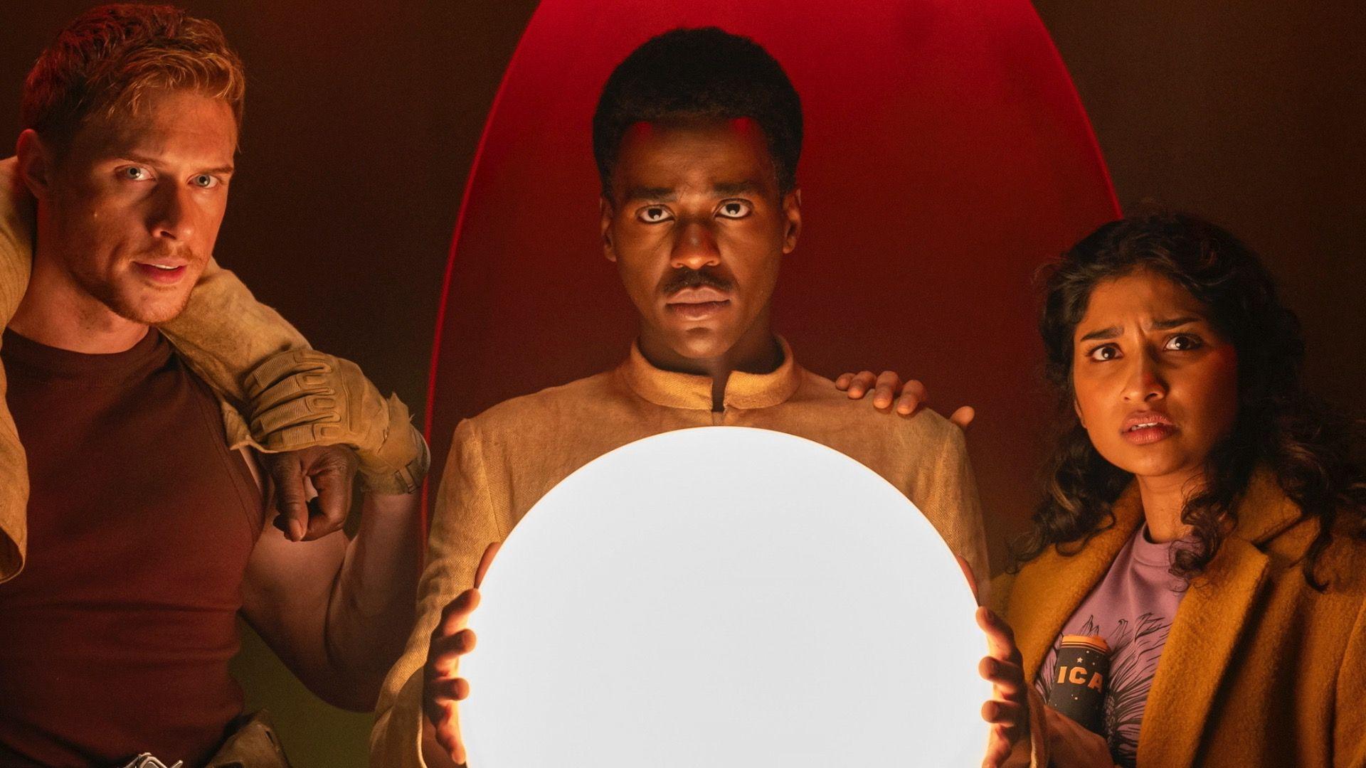The Doctor stands between Manny and Belinda as he holds a large glowing orb while staring into the camera
