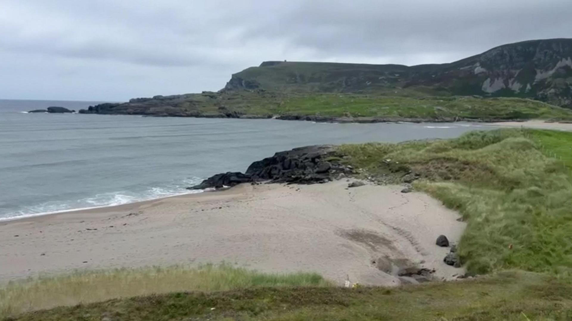 A small beach with light brown sand is in the foreground. There are small waves breaking on the shoreline and the sea is a slate gray colour. There are small grassy dunes to the right of the beach and a high headland in the background.