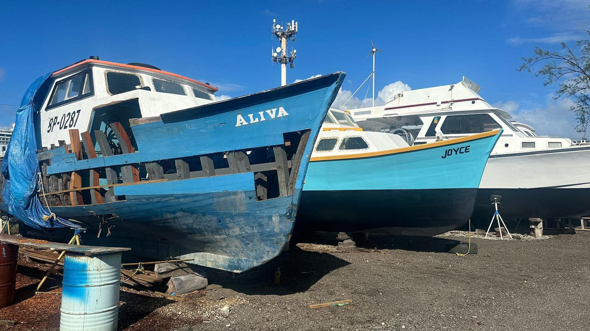 Damaged boat in dry dock with the name Aliva painted on the side, next to another repaired boat with the name Joyce on the side. The boats are blue and white against a bright blue sky.