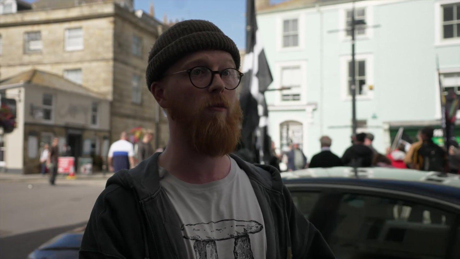 Sam Carmichael looking to the right as he talks to the reporter. He is wearing a black hat, a black hooded jacket and a white t-shirt. He has a beard and is wearing glasses. Behind him is two buildings with a road between them. There are people stood in front of the buildings. Just behind Sam is the top of a car.
