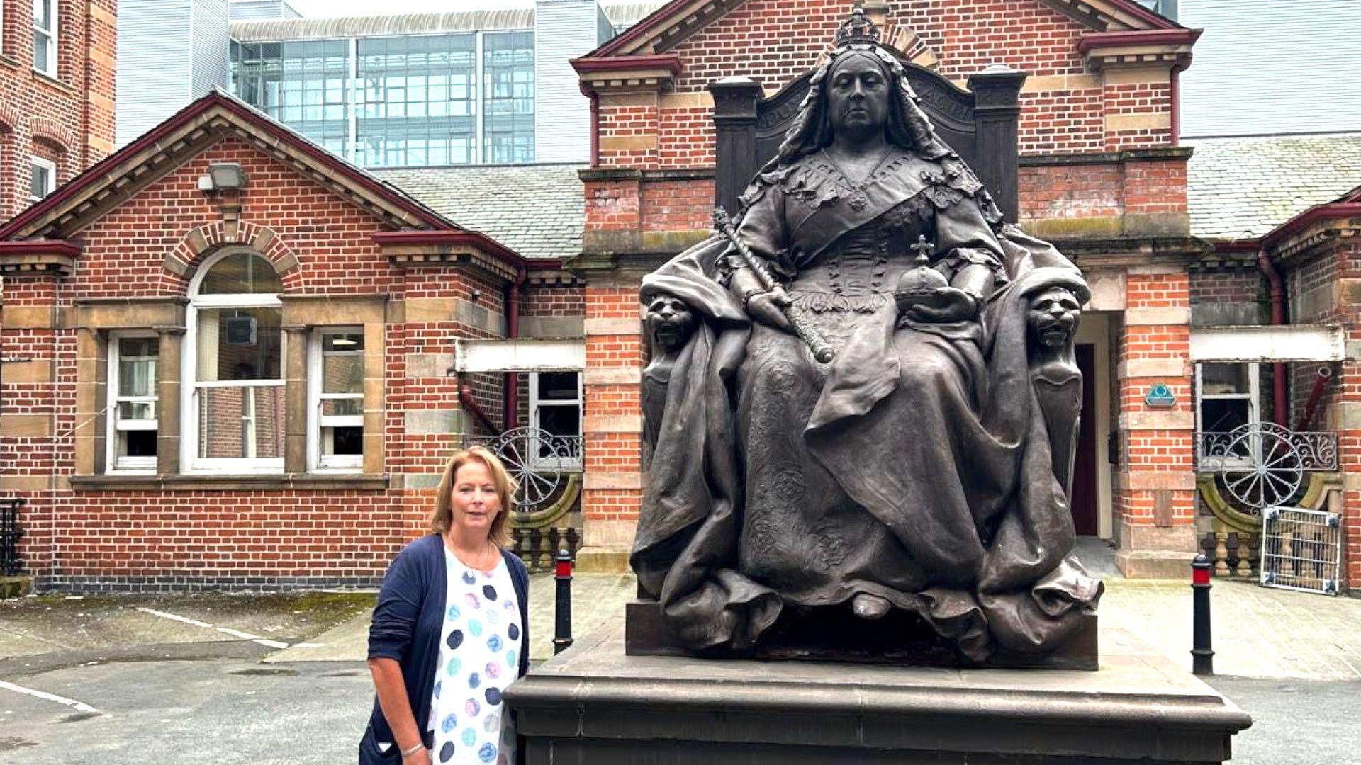 Monica pictured on the grounds of the Royal Victoria Hospital where she worked until her mesothelioma diagnosis. She is standing beside a large bronze statue of Queen Victoria sitting on a throne. Behind her are red-brick buildings of the older part of the hospital.