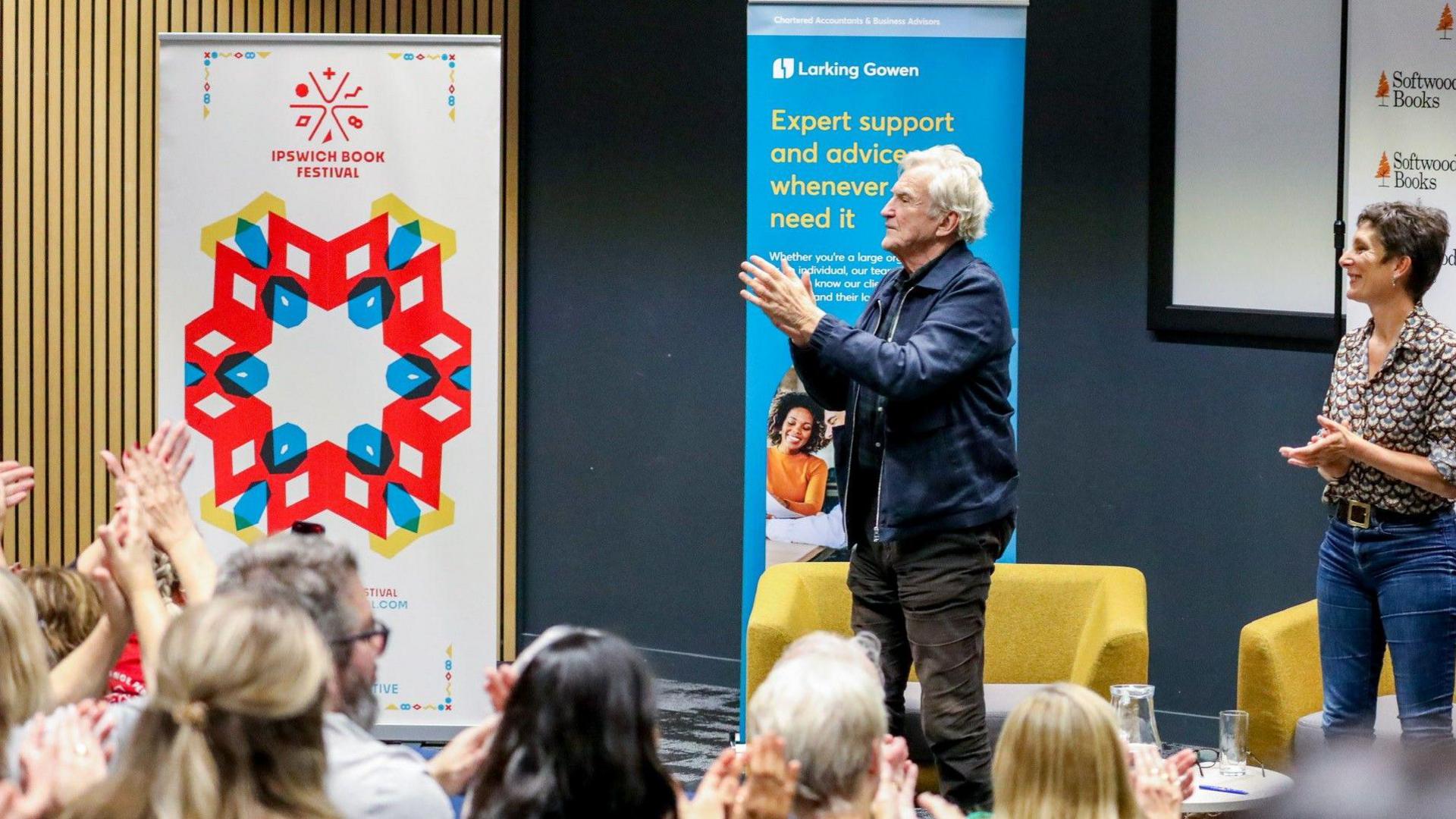Larry Lamb and Camilla Barnes stand on a stage and look to the left of the image. They are clapping. A crowd watches them from seats in front of them and are similarly clapping. 