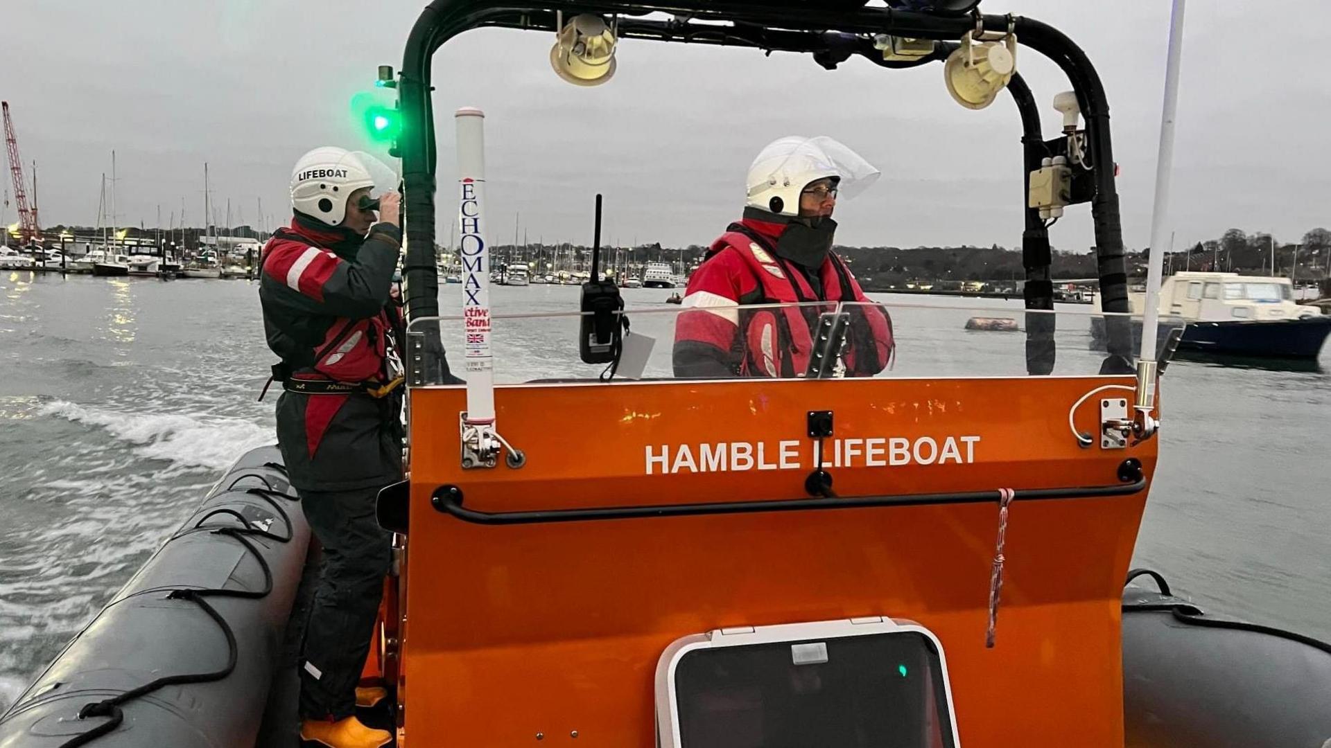 Two members of lifeboat crew stood on a black and orange boat. They are wearing red reflective suits with helmets and visors. One of the people is looking through a pair of binoculars.