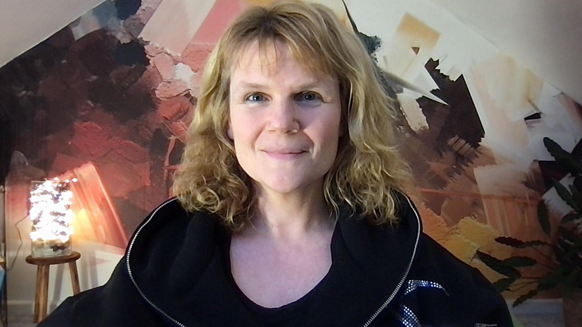 Claire Tchaikowski smiling, with blonde shoulder-lenght hair and wearing a black hoodie. 