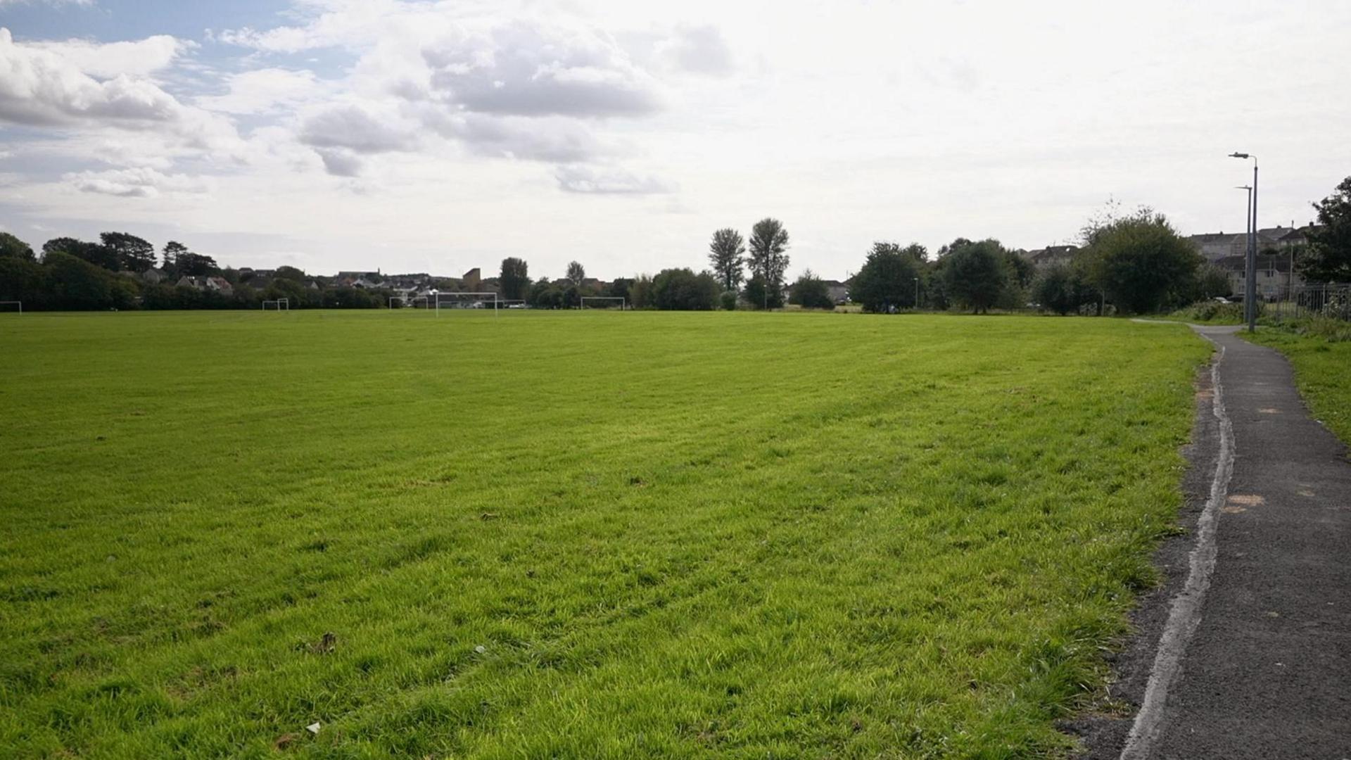 Playing fields made up of mostly grass, with a path on the right and trees, buildings and football posts in the distance