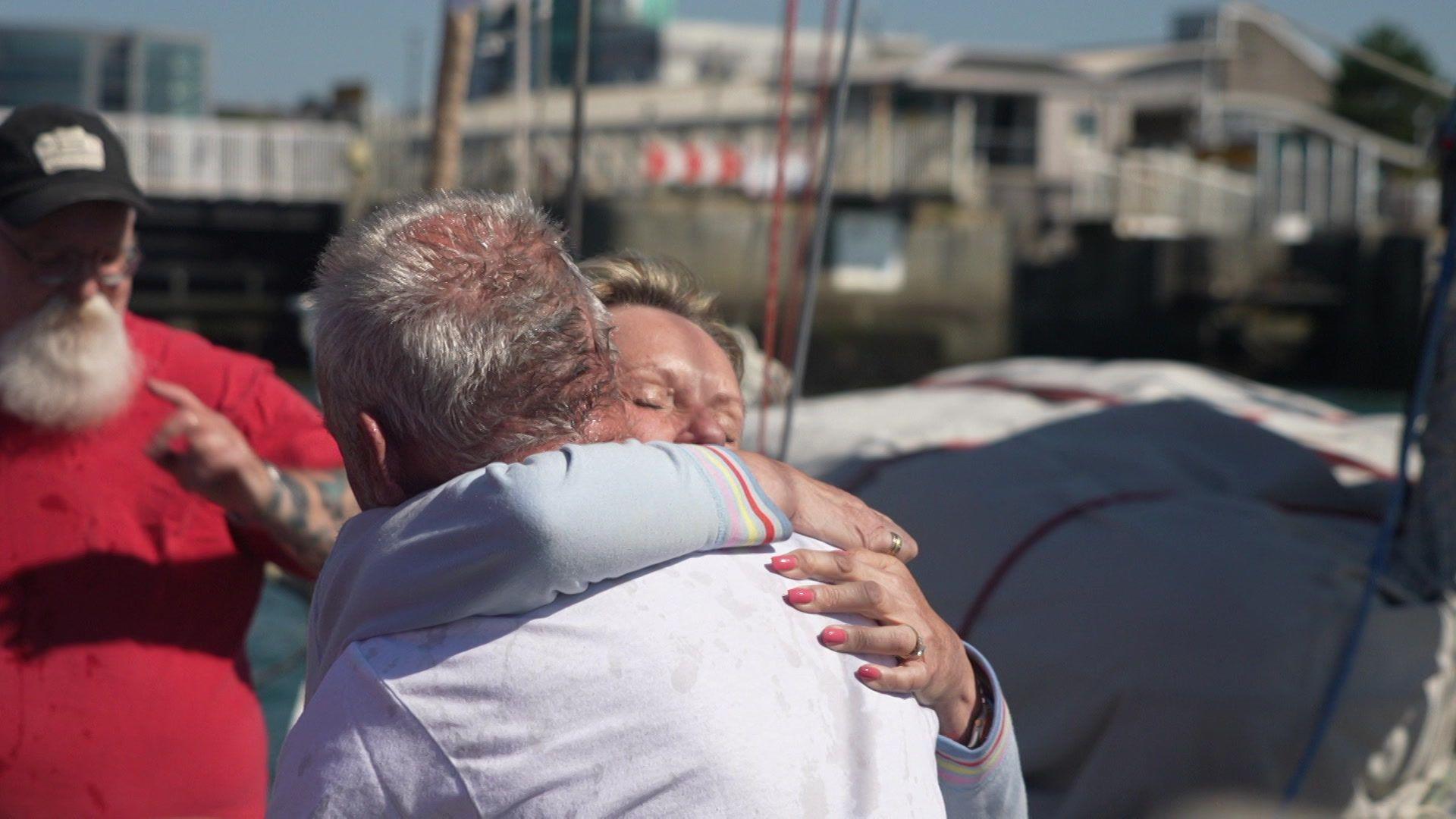 This picture shows three people near a waterfront. In the foreground, a person with gray hair wearing a white shirt is being hugged by another individual.
In the background, a third person with a white beard, wearing a red shirt and black cap, is gesturing or pointing.