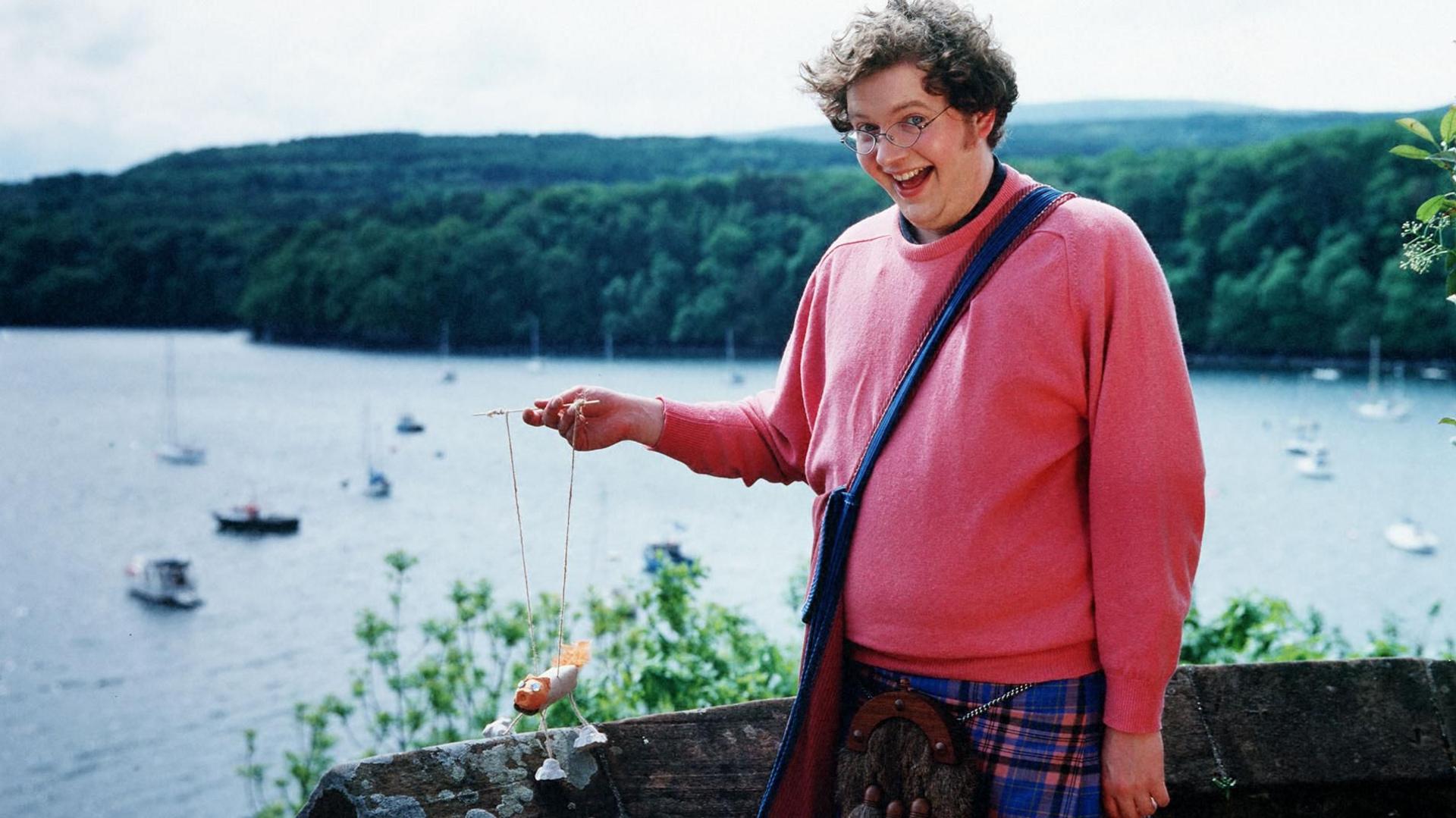 Miles Jupp, as Archie the inventor in Balamory, stands next to a low wall overlooking the harbour at Tobermory, holding a puppet made with egg boxes and wearing a pink jumper and kilt with sporran.