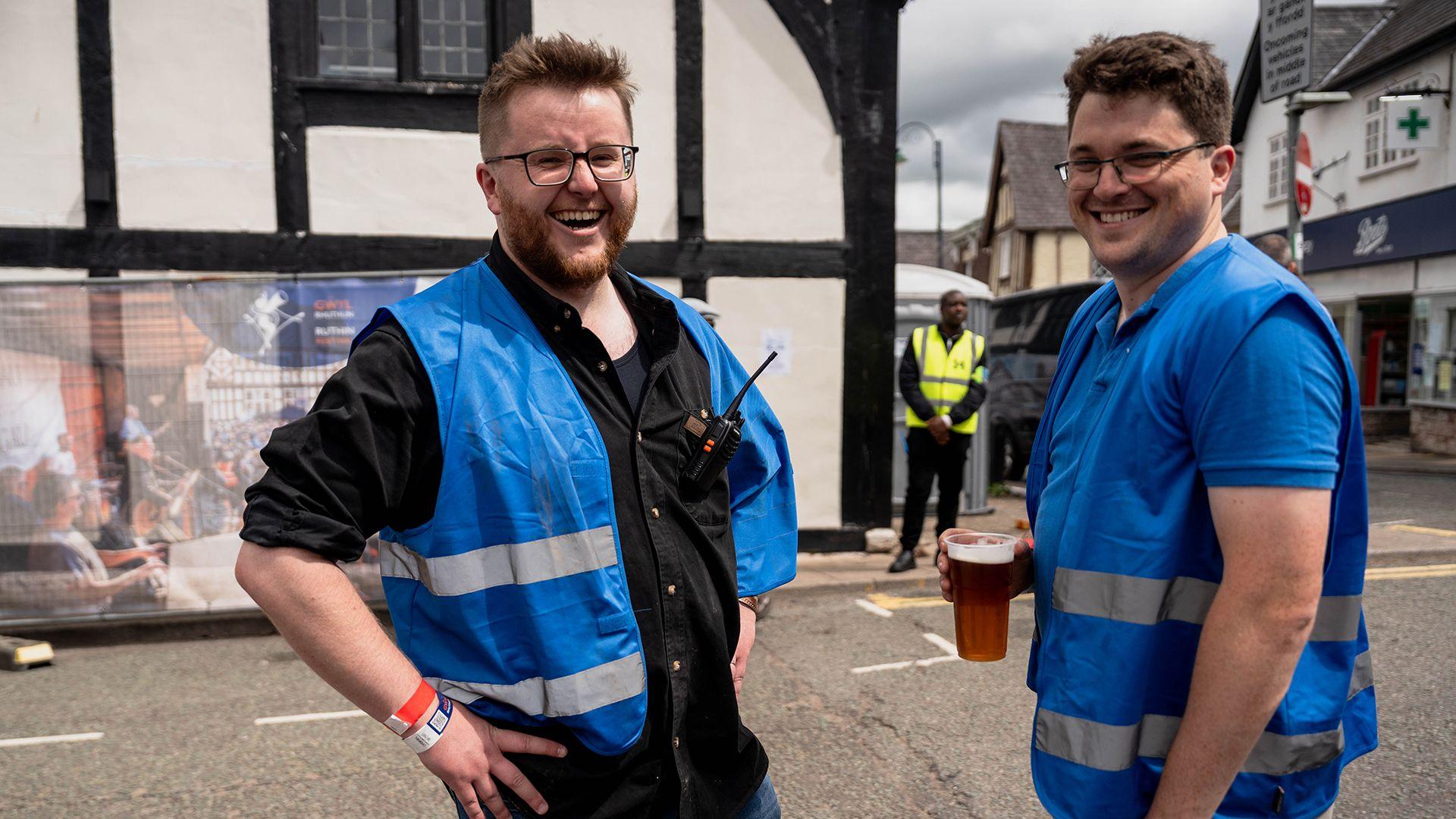 Dau ddyn efo 'high vis' yn gwenu, un yn cario peint