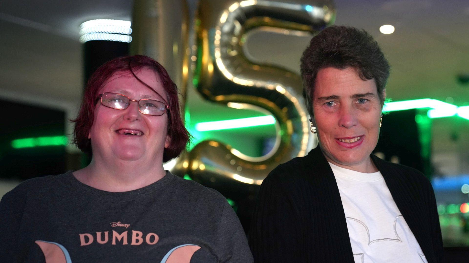 A woman wearing a grey "Dumbo" t-shirt stands on the left next to a woman wearing a black jacket and white 'Disney' t-shirt. They are in front of two large balloons in the shape of the numbers "1" and "5".