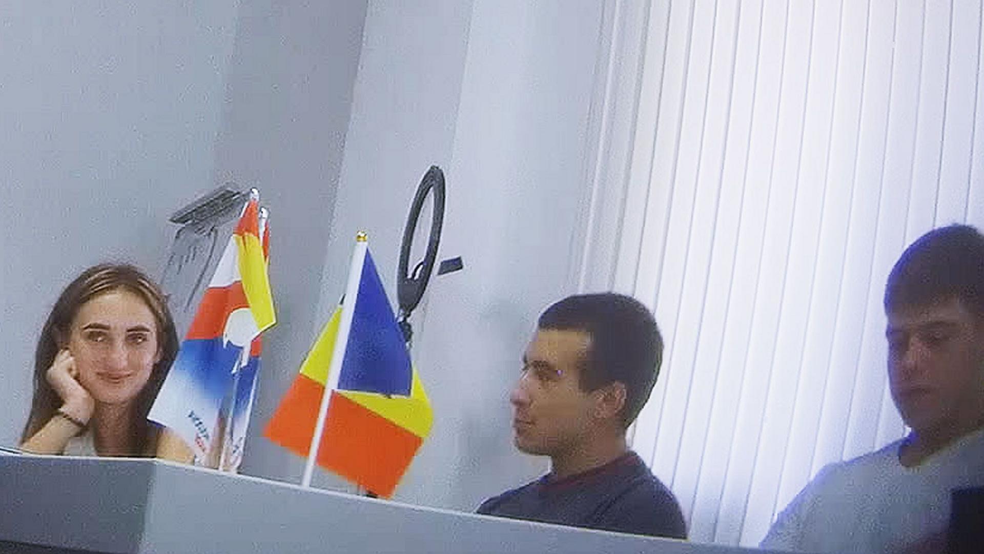 A woman with long brown hair (Alina Juk) smiles at the group surrounding her - they are sat around a large table and two flags are in view, one of which is the Moldovan flag in colours of red, yellow and blue