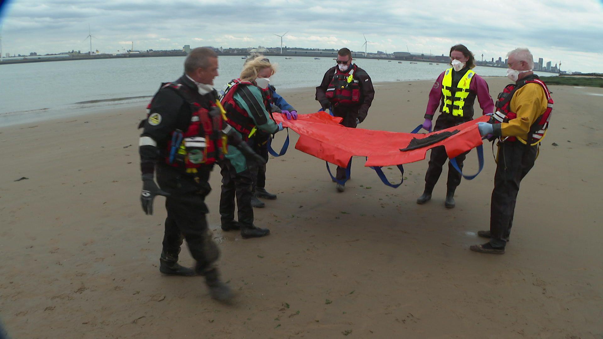 Six volunteers stand on the beach around a red stretcher with canvas straps