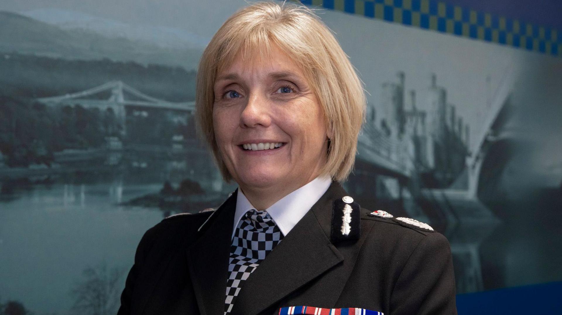 Amanda Blakeman is in a police uniform with a black and white checked cravat tie. She has badges on the shoulders of her black jacket. She has short blonde hair and blue eyes